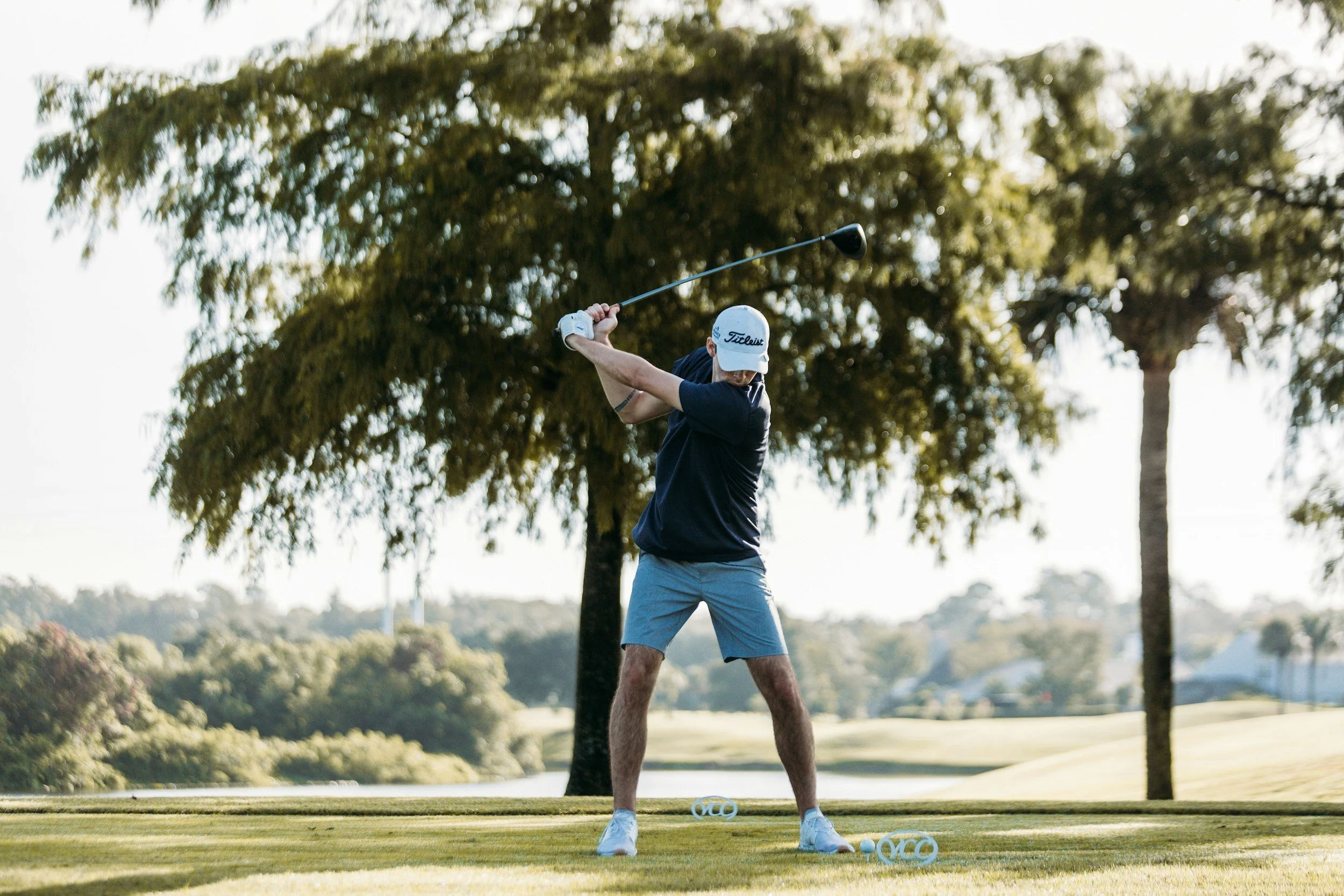 Man playing golf on a golf course, swinging a golf club, with trees and a pond in the background.