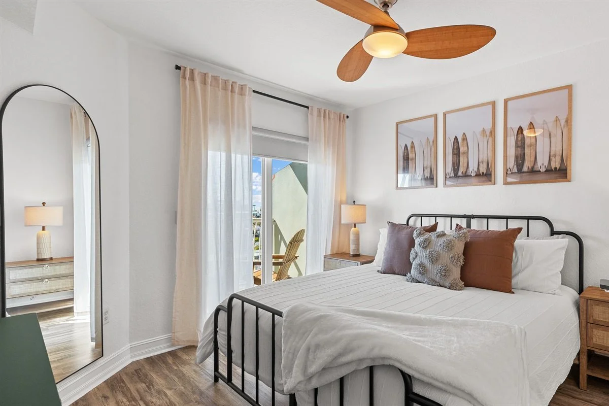 Bedroom with white walls, framed artwork, black metal bed frame, white bedding, and decorative pillows. Nightstands with lamps, a ceiling fan with wooden blades, a large mirror, and a balcony with an outdoor chair visible through glass doors.