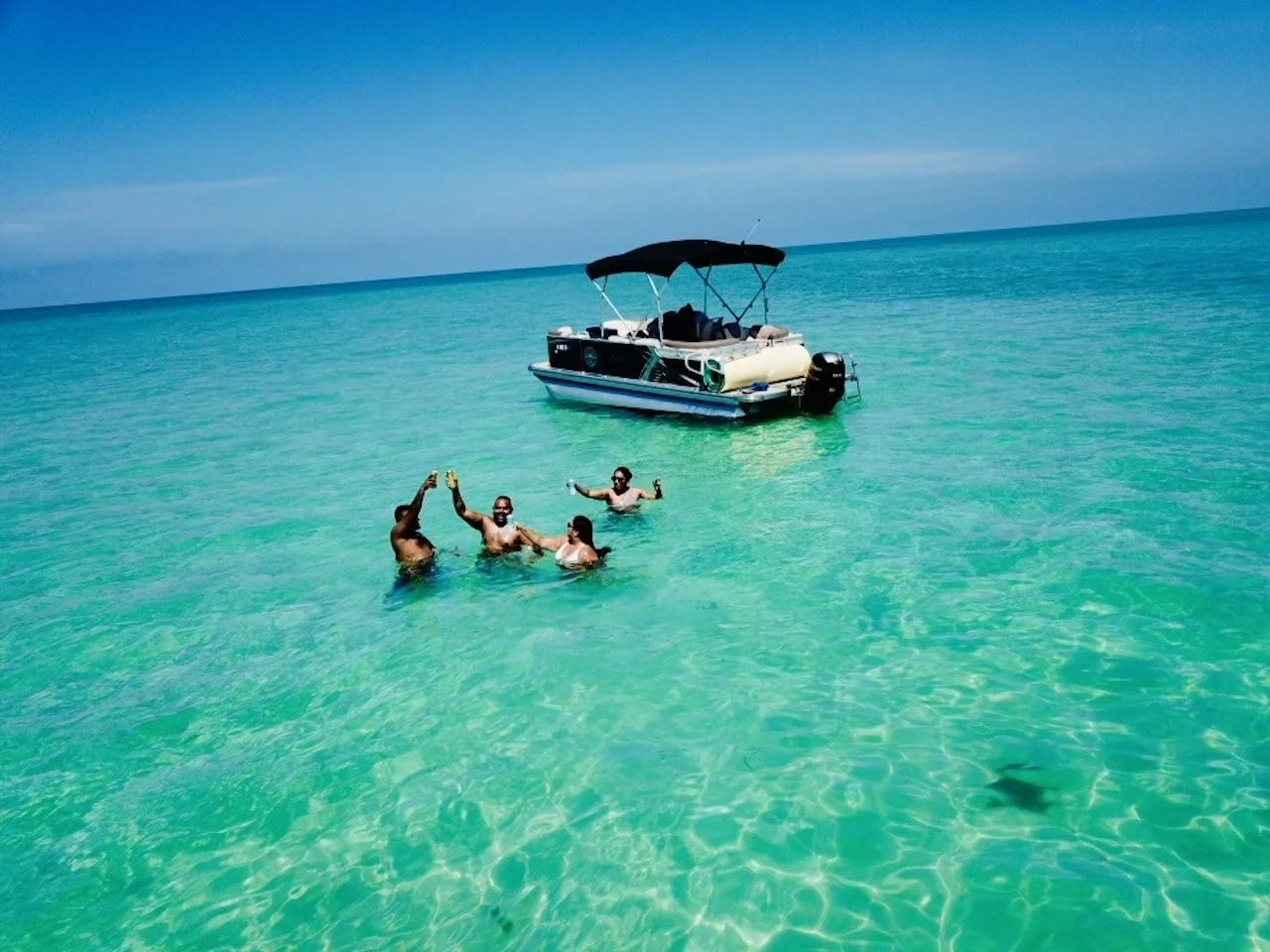 Four people swimming and celebrating in clear turquoise ocean water near a boat with a canopy.