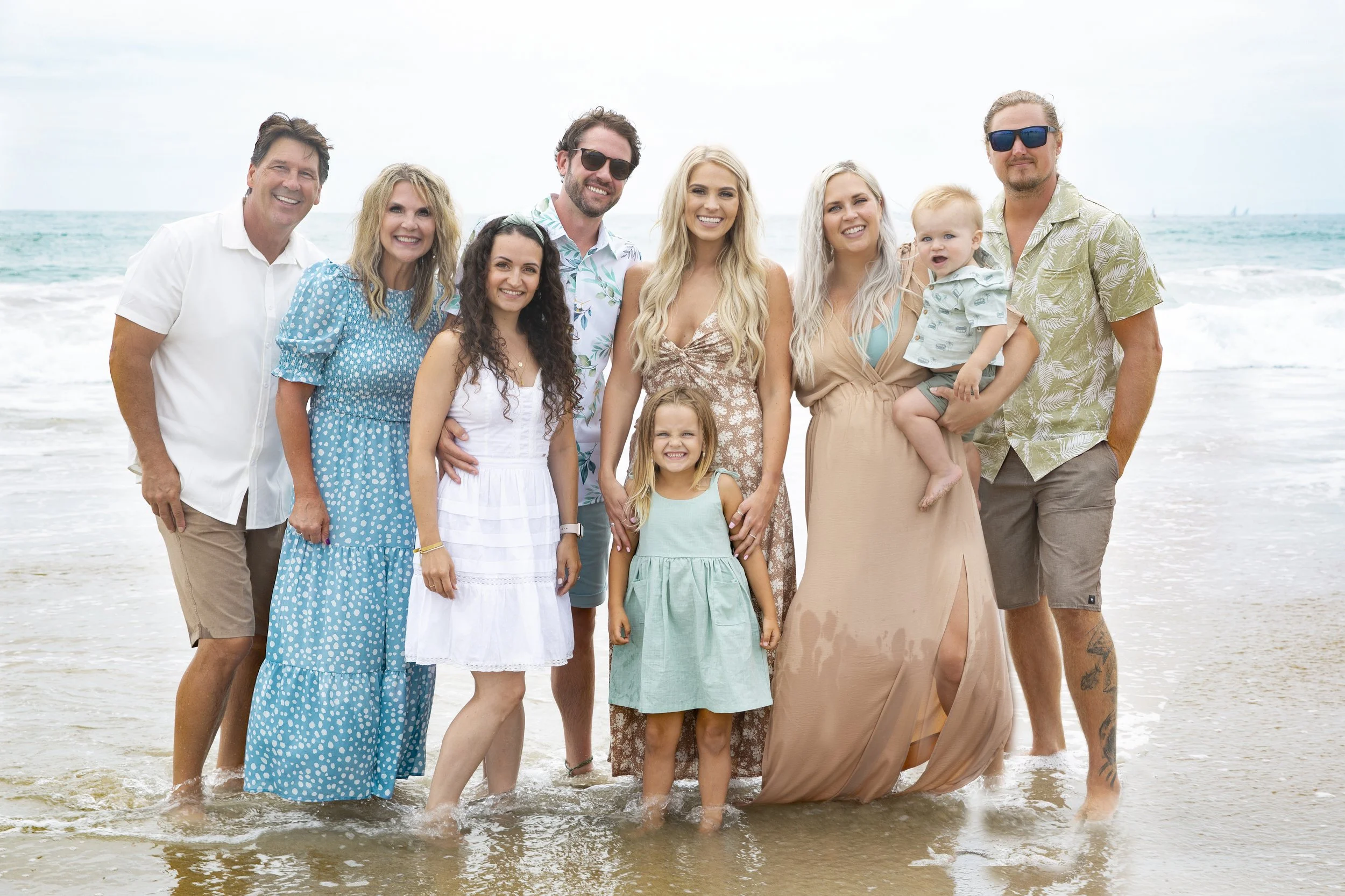 A group of ten people, including adults and children, smiling on the beach with the ocean and sky in the background.