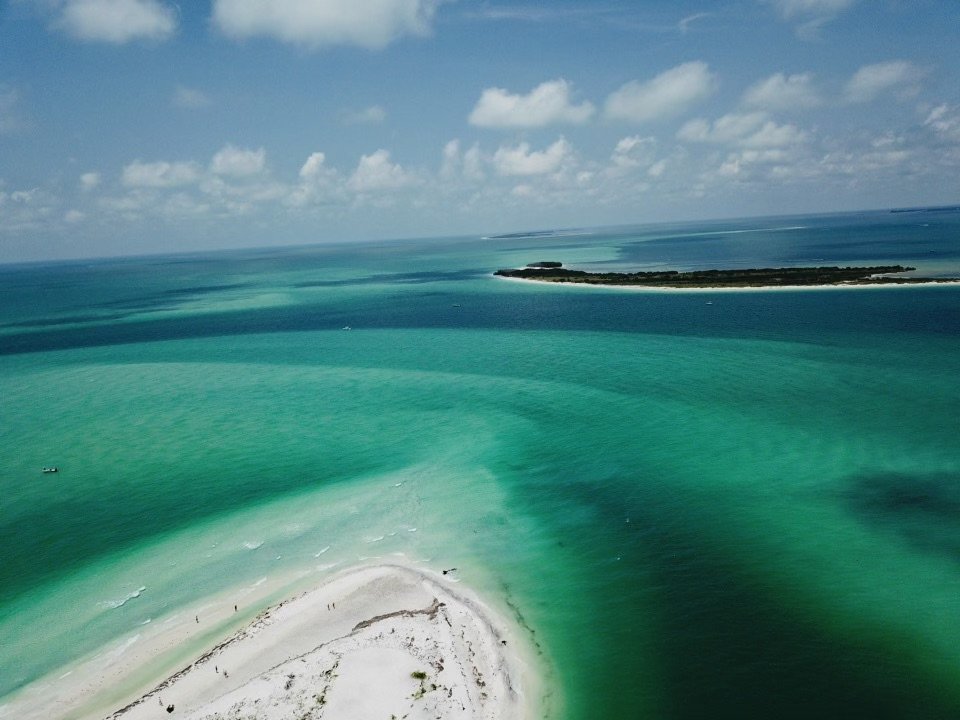 Aerial view of a tropical beach with white sand, turquoise water, and a small island in the distance under a partly cloudy sky.