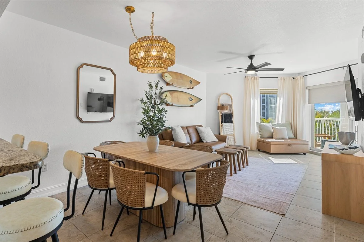 Bright living room and dining area with white walls, wicker pendant light, wall mirror, two fish wall decor, outdoor view, and tropical decor.
