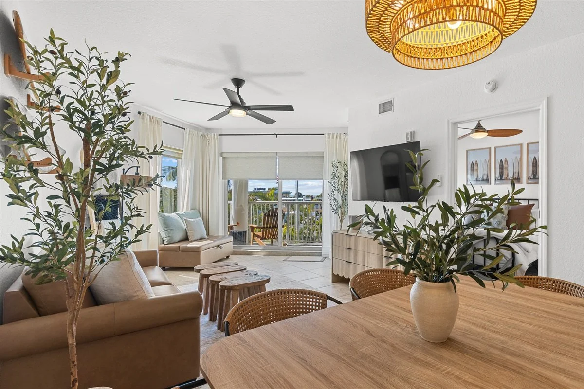 Living room with beige sofas, a wooden dining table with a vase of greenery, a mounted flat-screen TV, large windows with white curtains, and outdoor balcony with chairs.