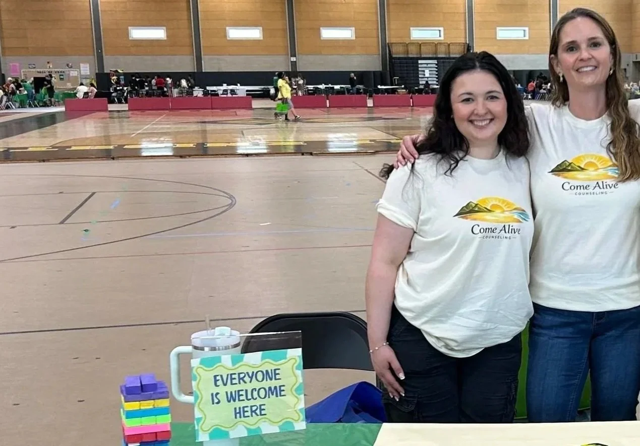 Two women wearing Come Alive Counseling t-shirts standing in front of a table with a sign that says 'Everyone is Welcome Here' inside a gymnasium or indoor sports facility with many people and booths in the background.