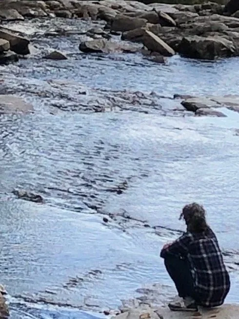 A person sitting on rocks by a river, gazing at the water.