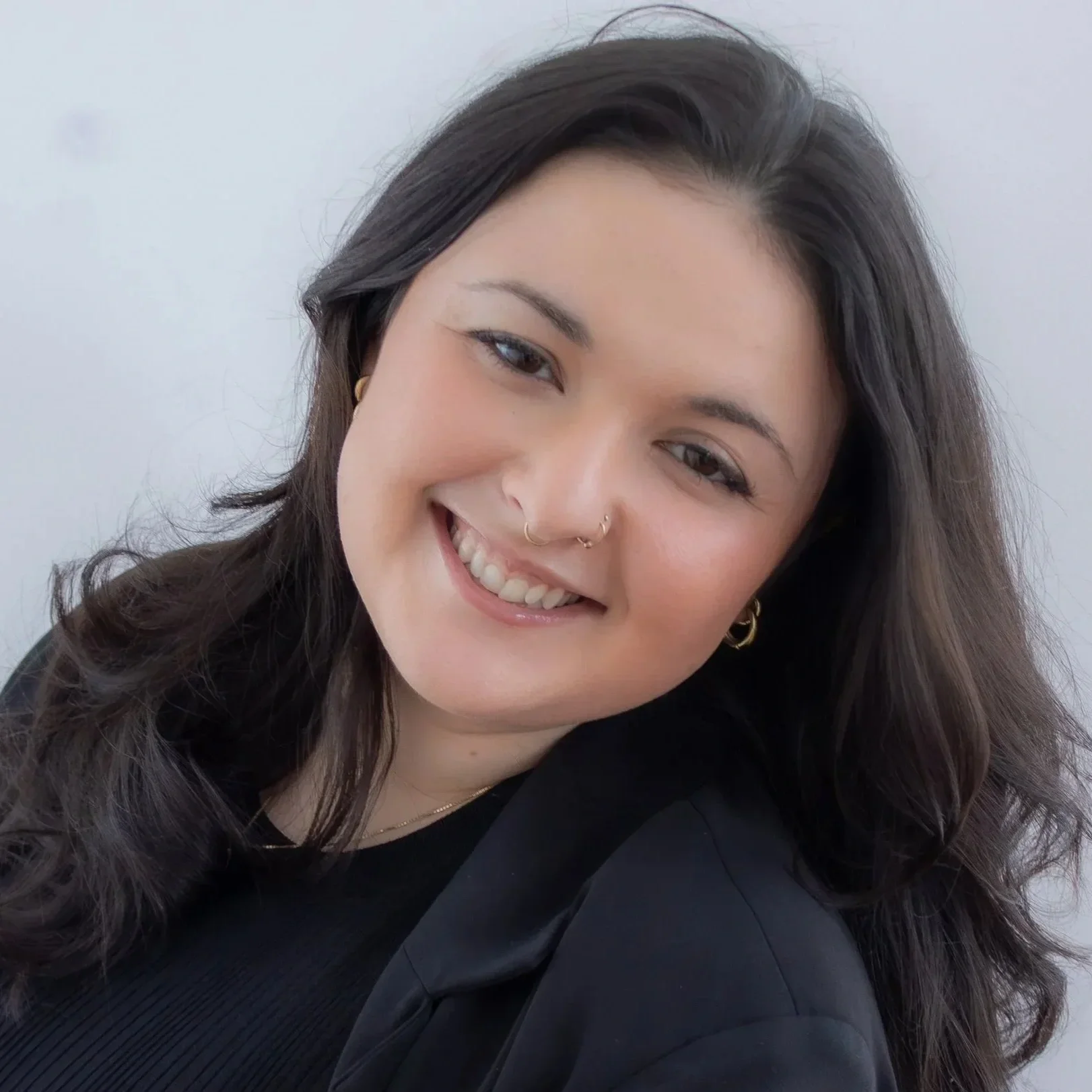 A woman with long wavy dark hair smiling, wearing gold hoop earrings and a nose ring, dressed in black, with a white background.