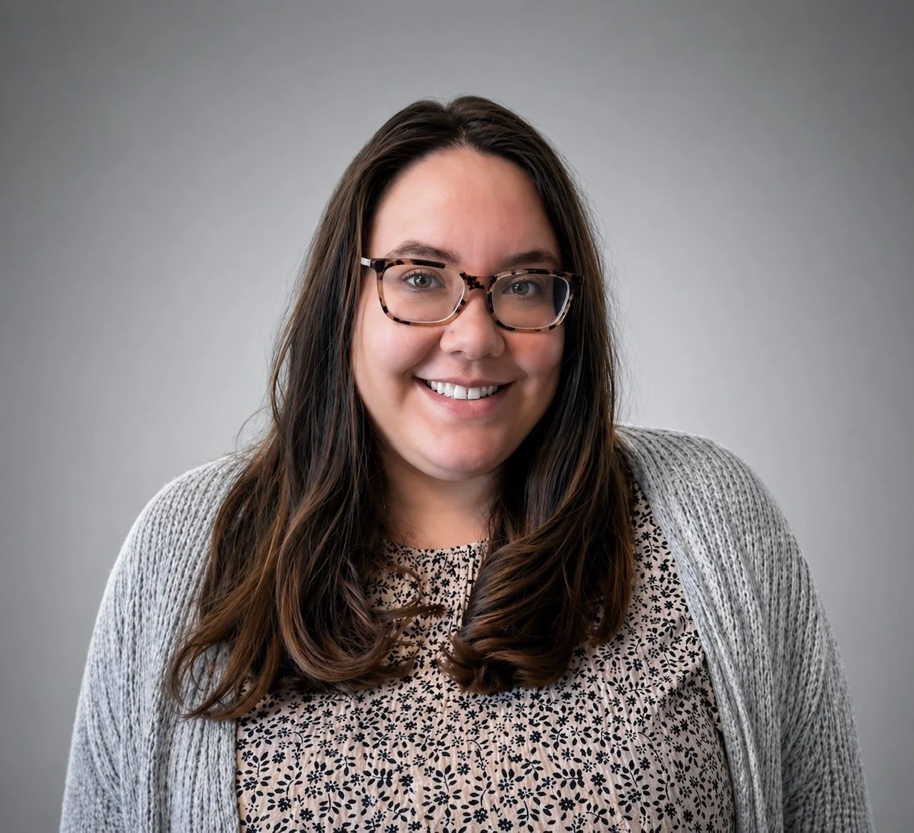 A smiling woman with long brown hair, glasses, a patterned shirt, and a light gray cardigan standing against a plain gray background.