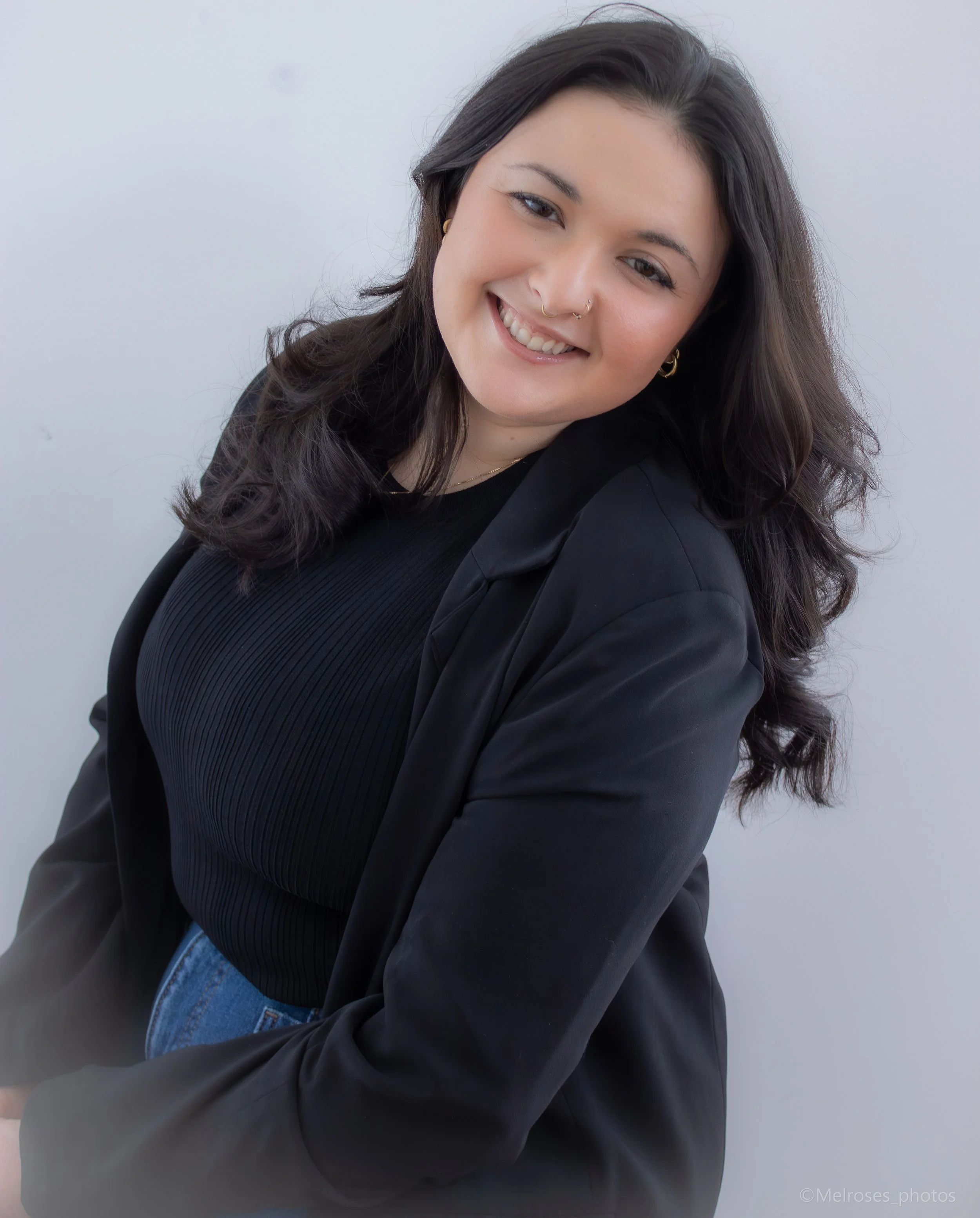 A young woman with dark wavy hair, smiling in a black blazer and black top, against a light gray background.