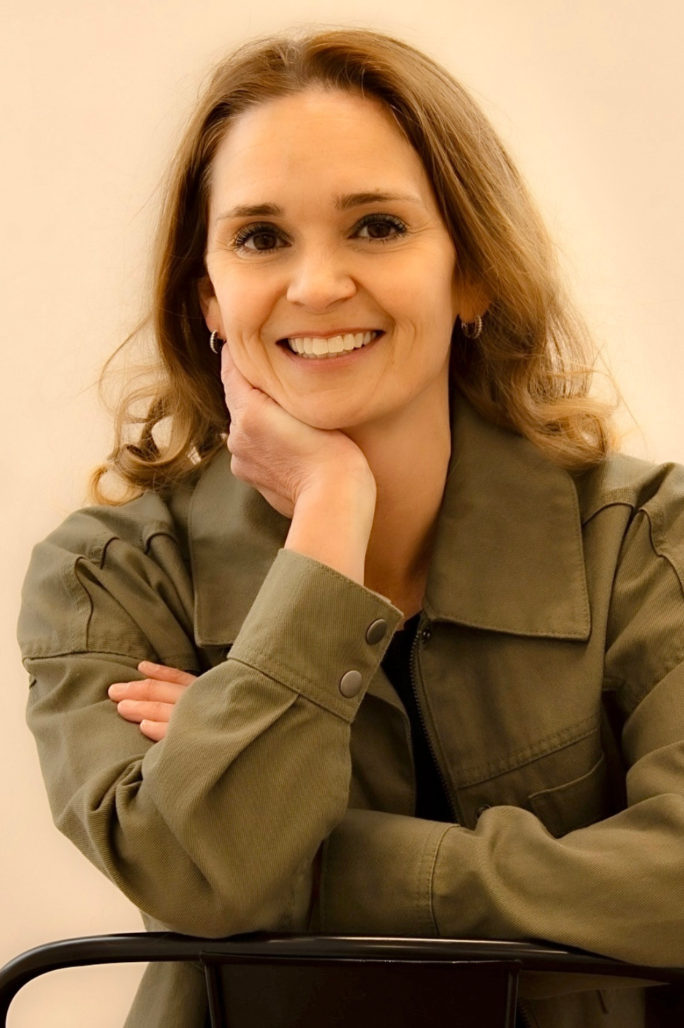 A woman with light brown hair, smiling, resting her chin on her hand. She is wearing a green jacket and seated against a plain light-colored background.