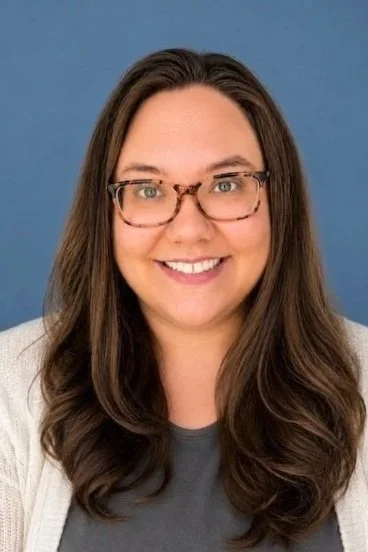 A woman with long, wavy brown hair wearing glasses, a gray shirt, and a light-colored cardigan, smiling in front of a blue background.