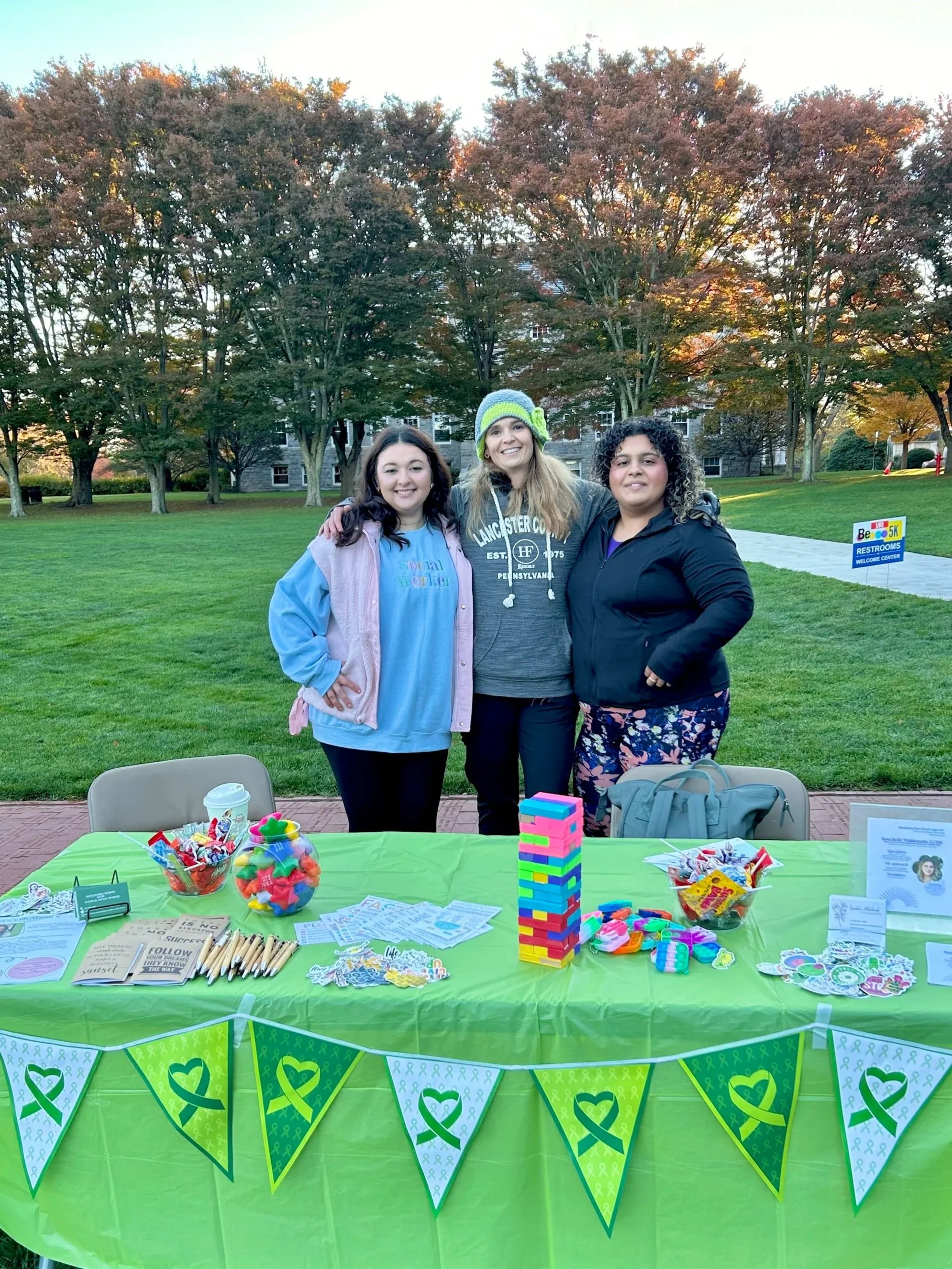 Three women standing behind a table with colorful candy and game pieces, outdoors in a park with trees in the background.