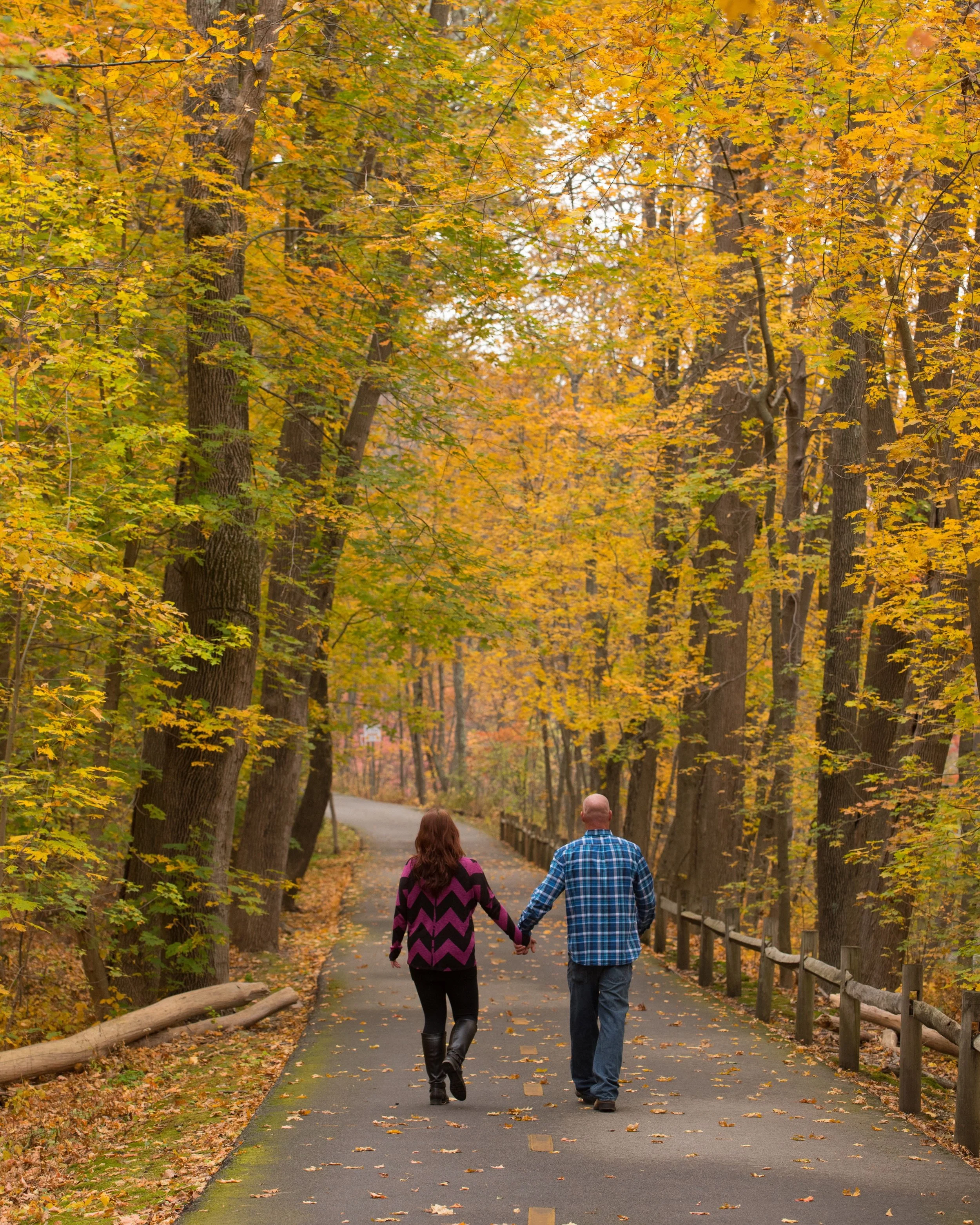 A couple walks hand-in-hand down a paved, tree-lined path through a forest in autumn, with leaves in shades of yellow, orange, and green, and fallen leaves on the ground.