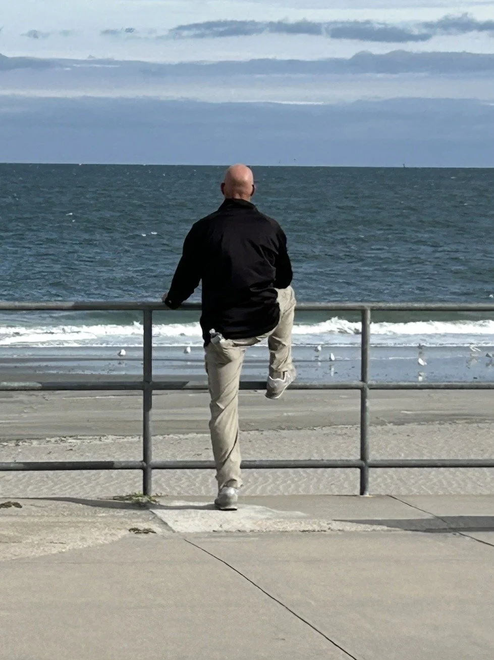 A man with a bald head, wearing a black jacket and beige pants, stands by a metal railing, looking out at the ocean on a partly cloudy day.