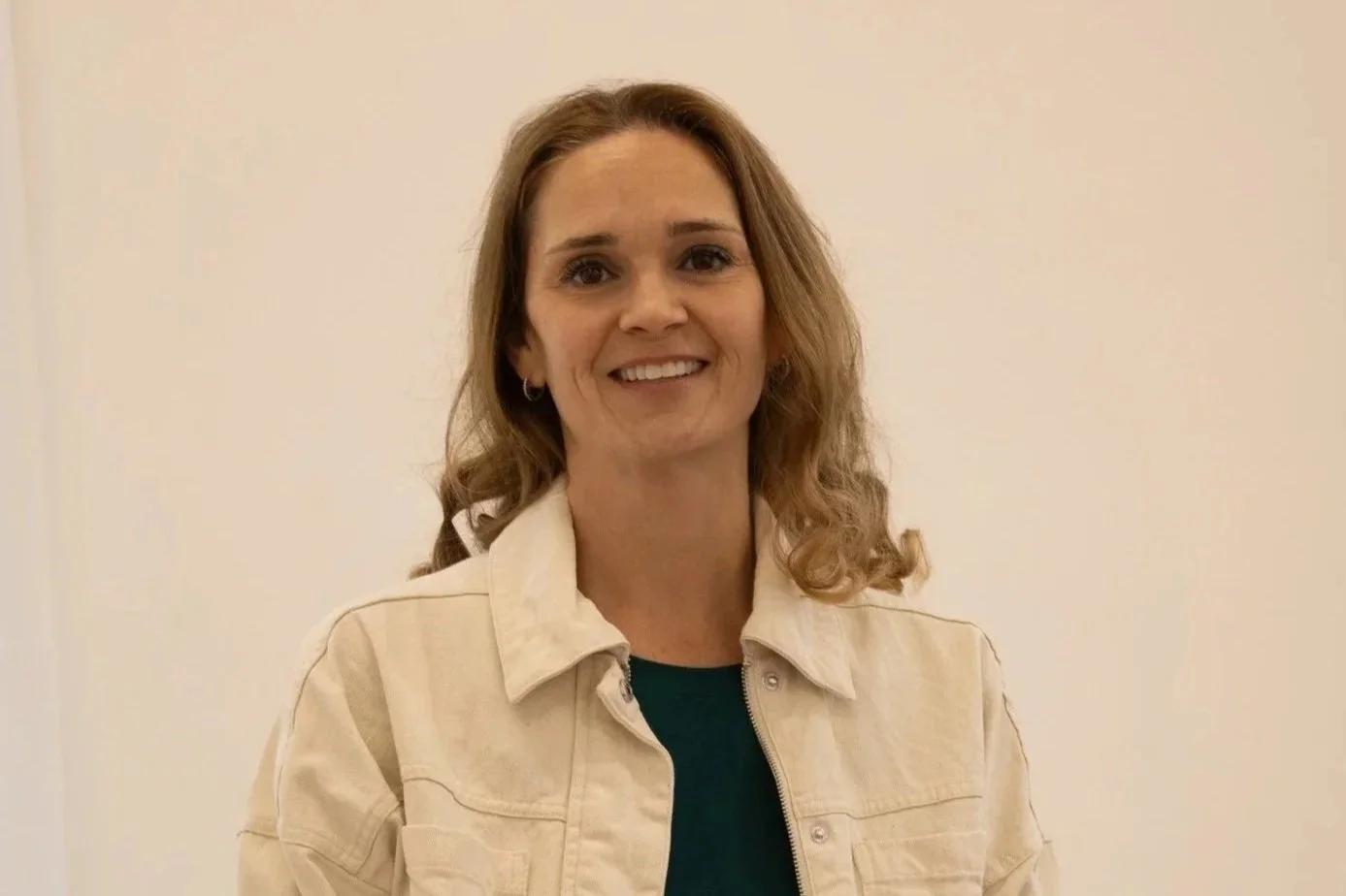 A woman with wavy brown hair smiling, wearing a beige jacket over a dark green top, standing in front of a plain off-white wall.