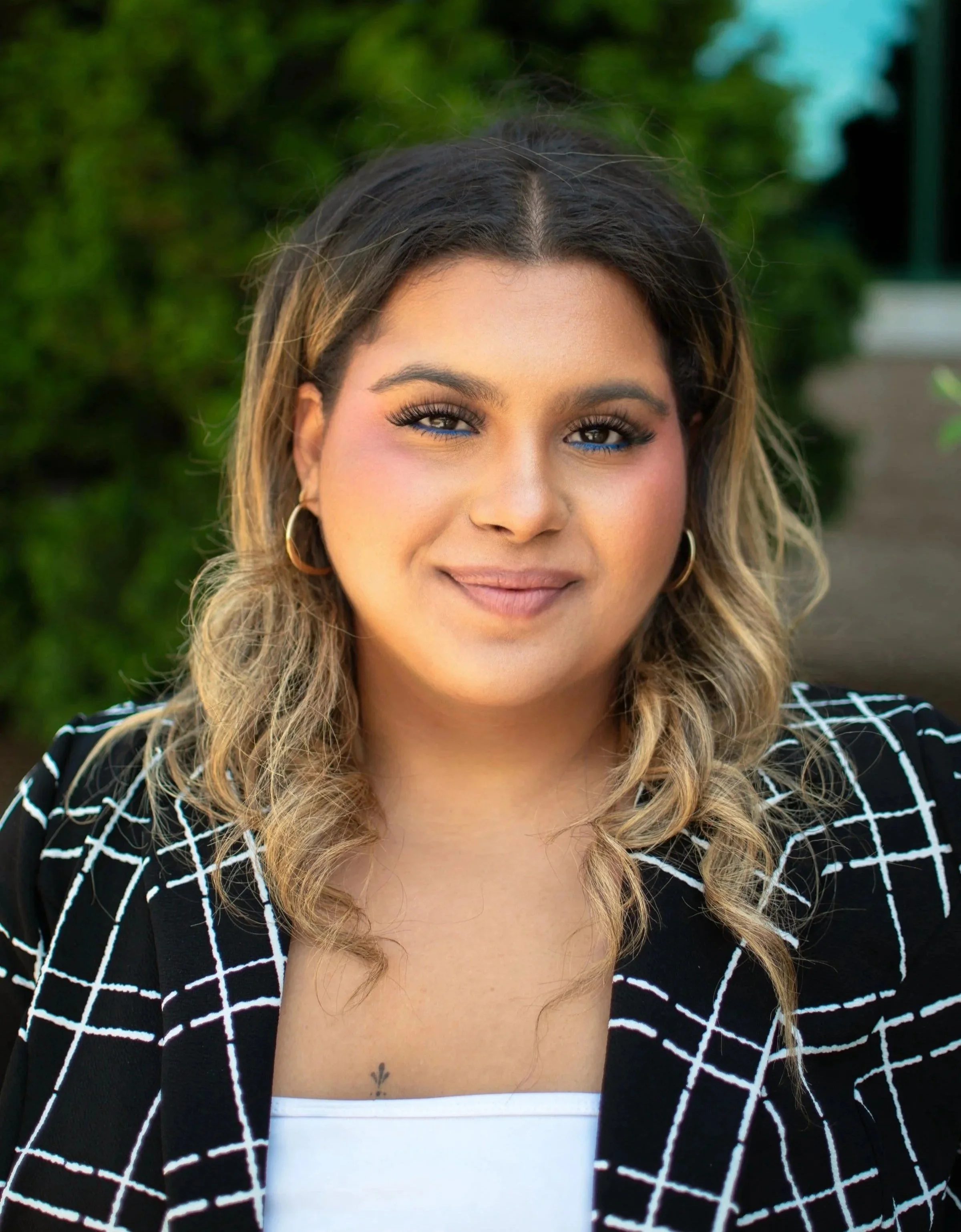 A woman with light brown hair and golden highlights, wearing a black and white checkered blazer, smiling outdoors with greenery in the background.