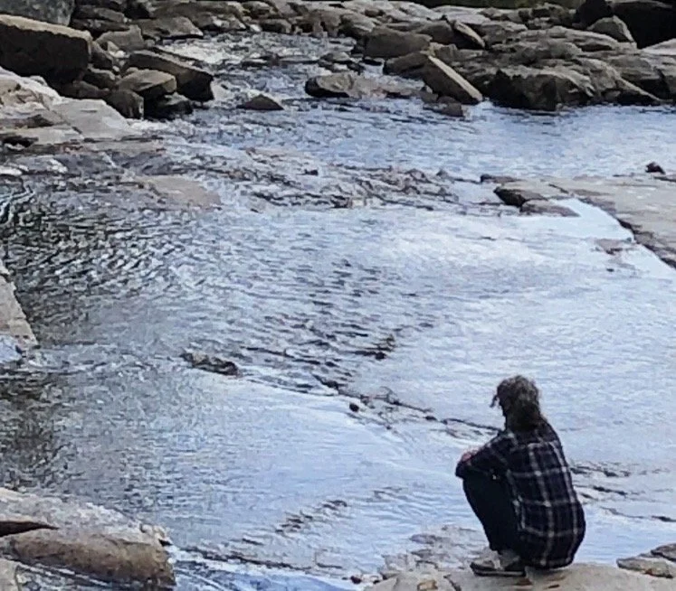 A person sitting on rocks next to a river or stream, surrounded by larger rocks and flowing water.