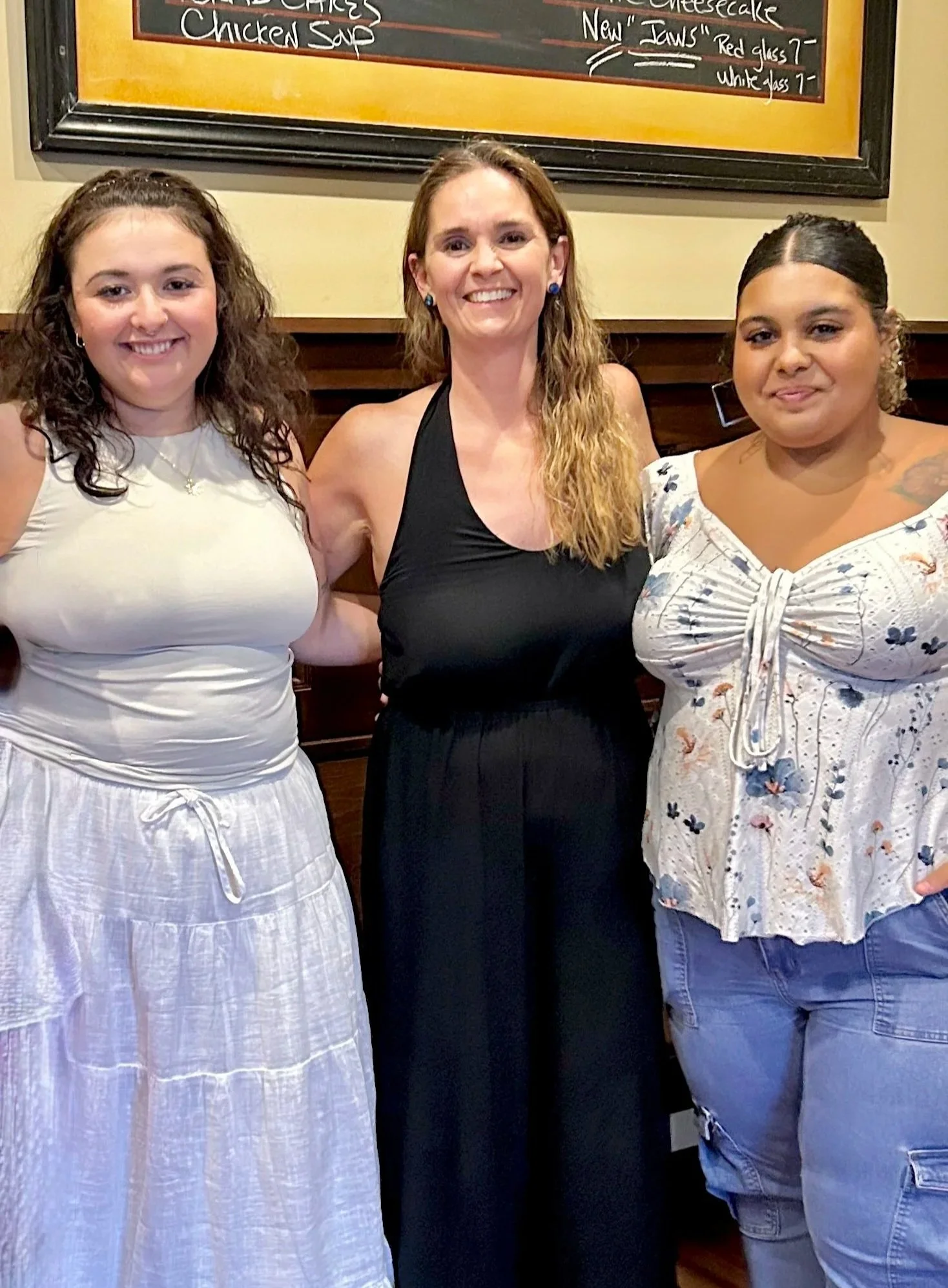 Three women standing side by side in a restaurant with a chalkboard menu in the background. The woman on the left has curly brown hair and is wearing a white dress. The woman in the middle has long wavy hair and is dressed in a black sleeveless top and a black skirt. The woman on the right has dark hair tied back and is wearing a floral top and blue jeans.