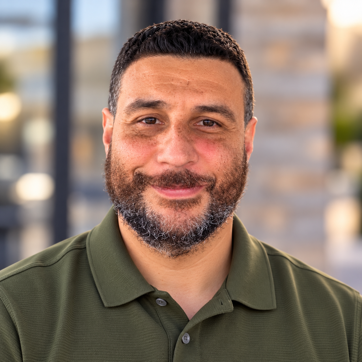 Close-up portrait of a smiling man with dark hair and a beard, wearing a green polo shirt. Blurred background with a modern building.