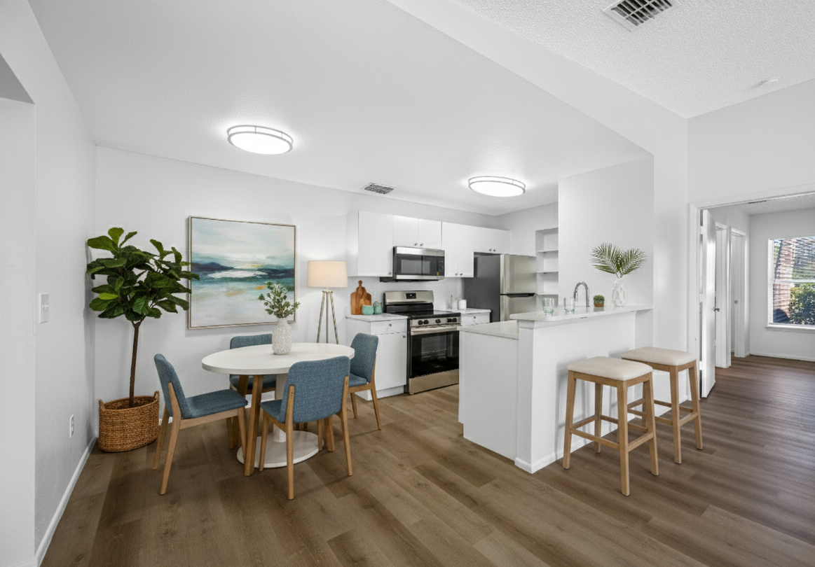 Open-concept kitchen and dining area with white walls, wooden flooring, and modern furniture. The dining table has four blue chairs, and a large potted plant is nearby. The kitchen features white cabinets, stainless steel appliances, and a breakfast bar with two stools. Decorative items and a painting are on the walls and counters, and there is natural light coming through a window.