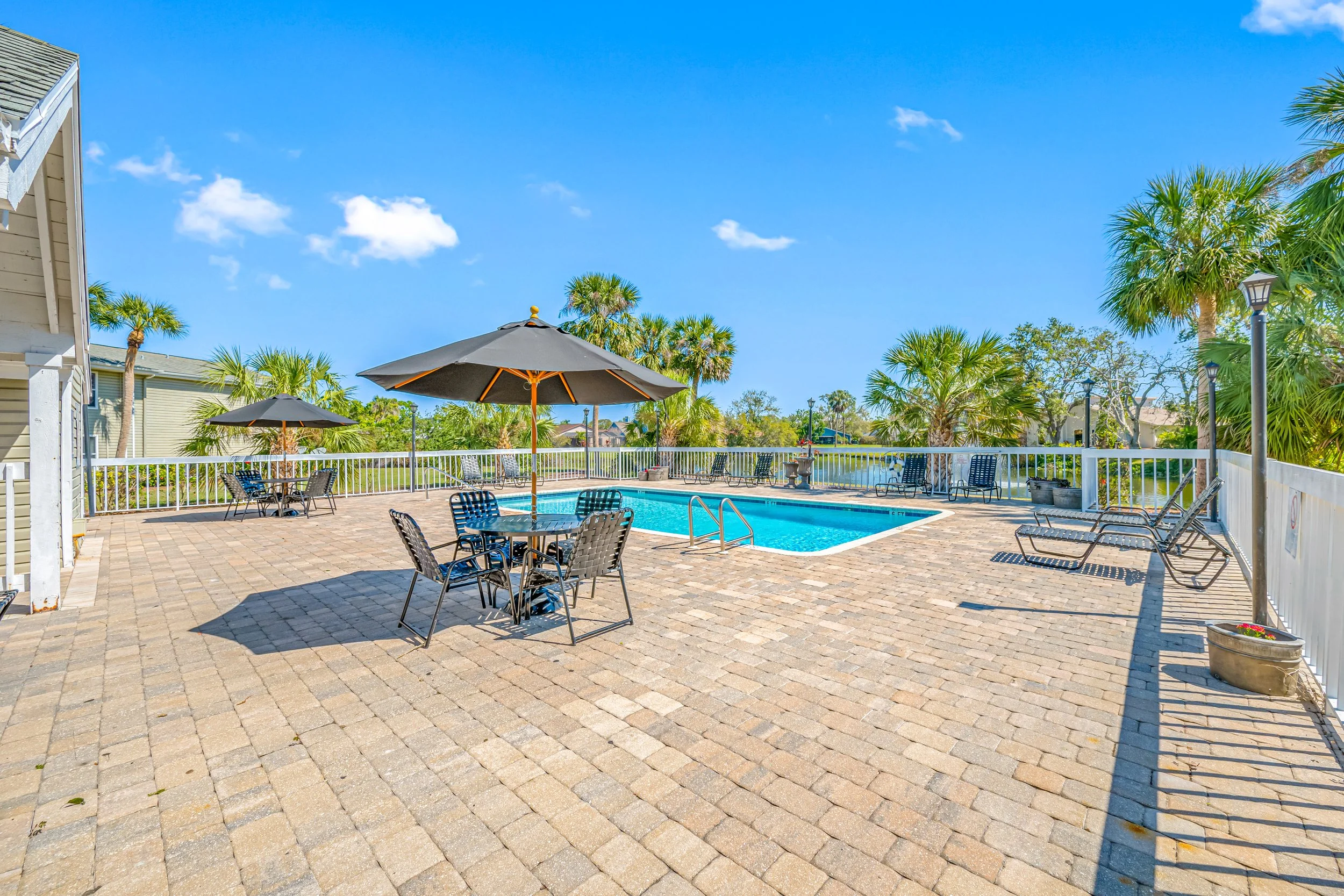 Swimming pool area with tables, chairs, umbrellas, and lounge chairs on a brick patio, surrounded by palm trees and a white fence under a clear blue sky.