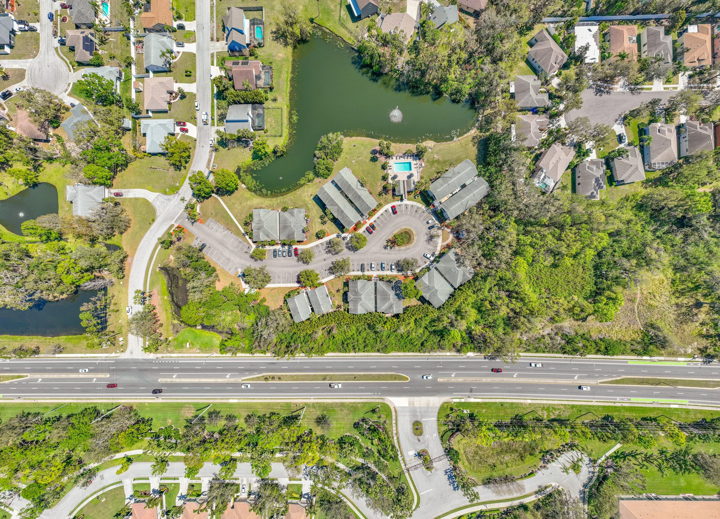 Aerial view of a suburban residential complex with multiple buildings, a parking lot, a small pond, and a larger pond with a fountain, surrounded by trees and roads.