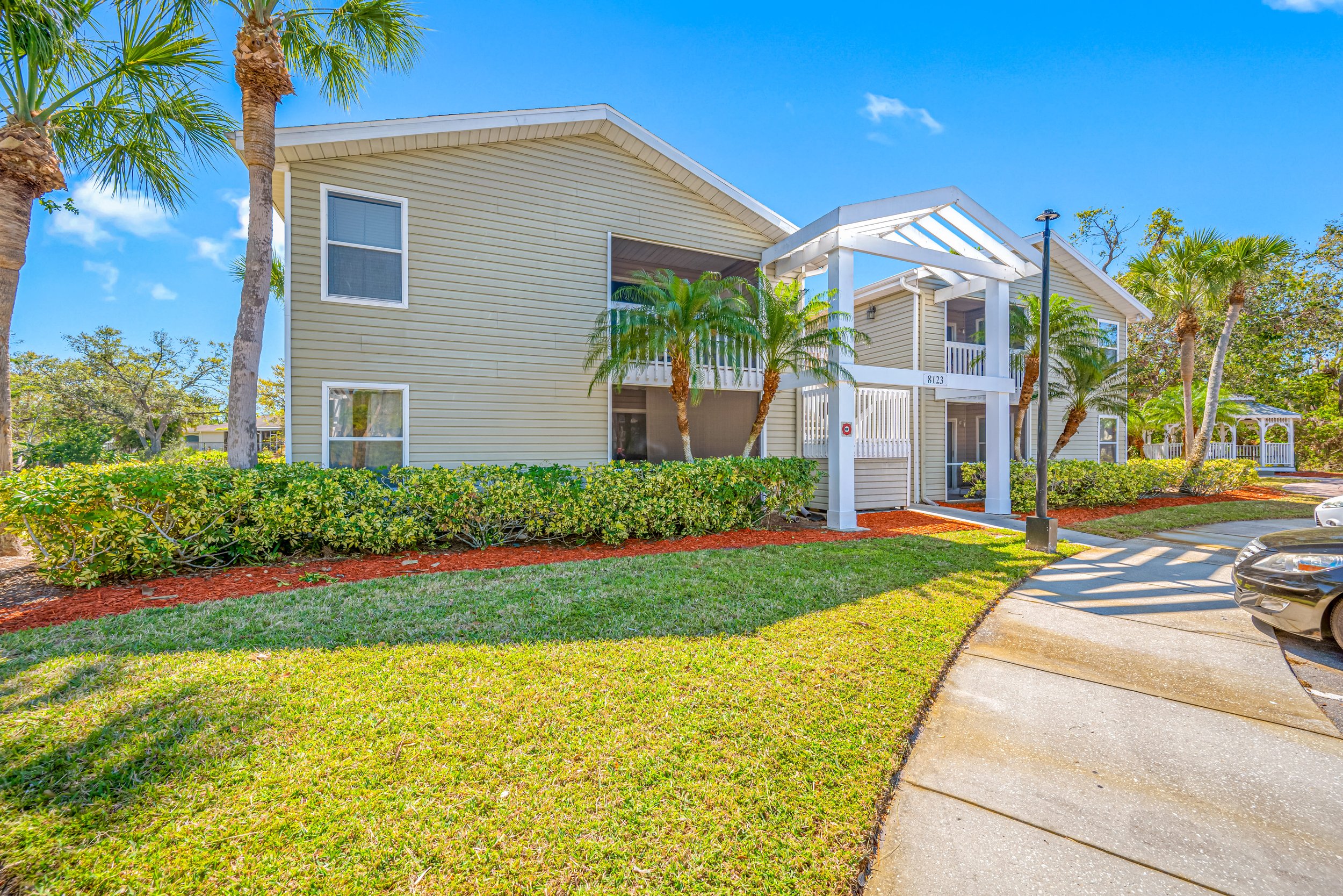 Exterior view of a two-story residential building with beige siding, surrounded by tropical palm trees, well-maintained grass and bushes, and a curved concrete driveway with parked cars.