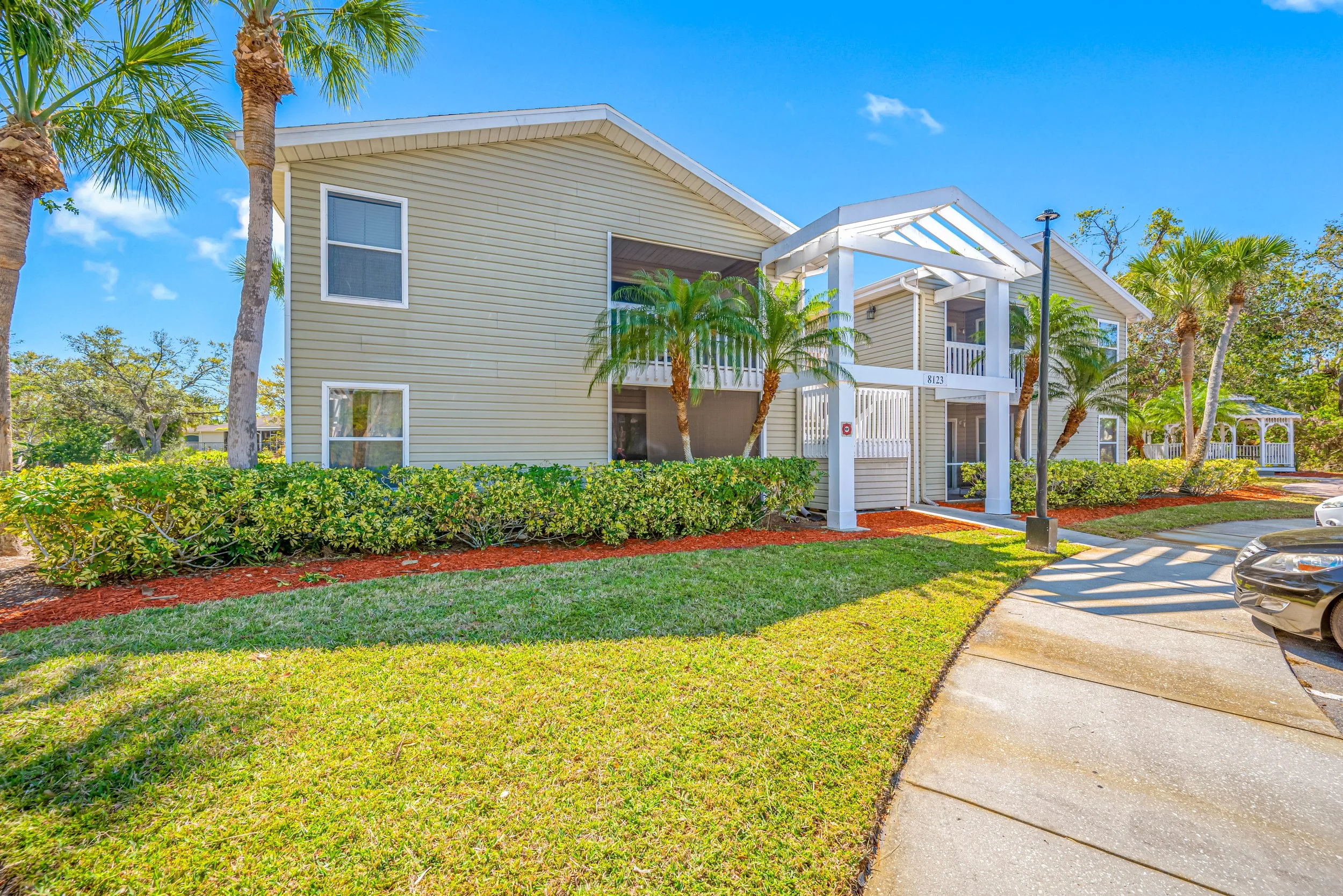 A two-story yellow apartment complex with white trim surrounded by green bushes and palm trees, with a clear blue sky overhead and a concrete sidewalk leading to the entrance.