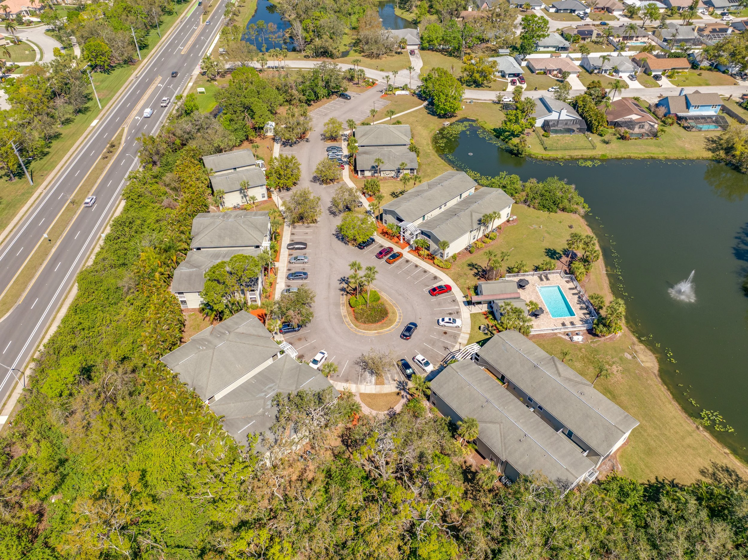 An aerial view of a residential complex with multiple buildings, a parking lot, a swimming pool, and surrounding trees, nearby a road with vehicle traffic and a pond with a fountain.
