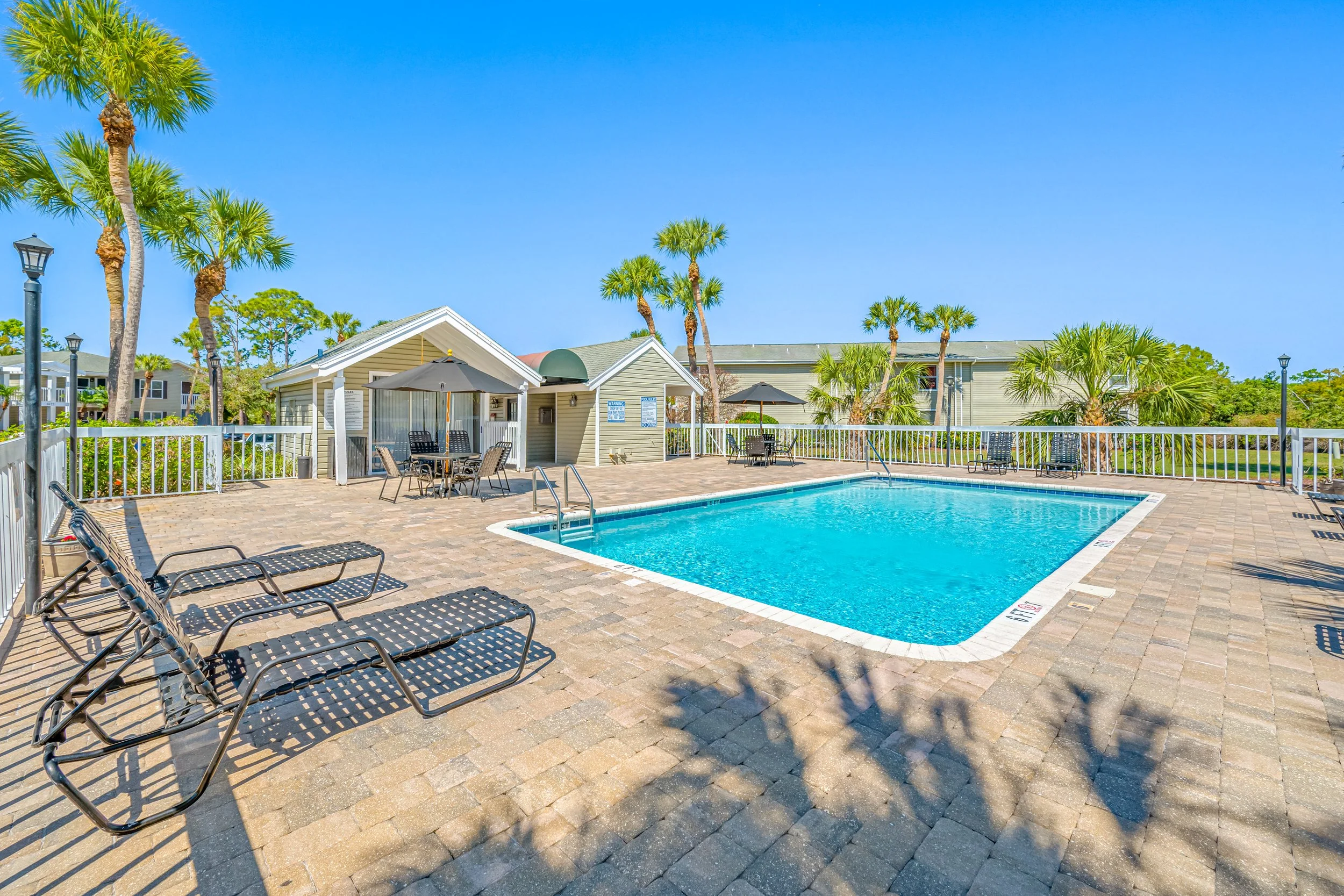 Empty outdoor swimming pool area with lounge chairs, tables with umbrellas, and palm trees under a clear blue sky.