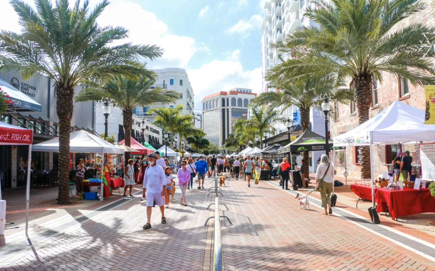 A lively outdoor market on a sunny day with palm trees, vendors' tents, and shoppers walking along a brick-paved street in a downtown area.