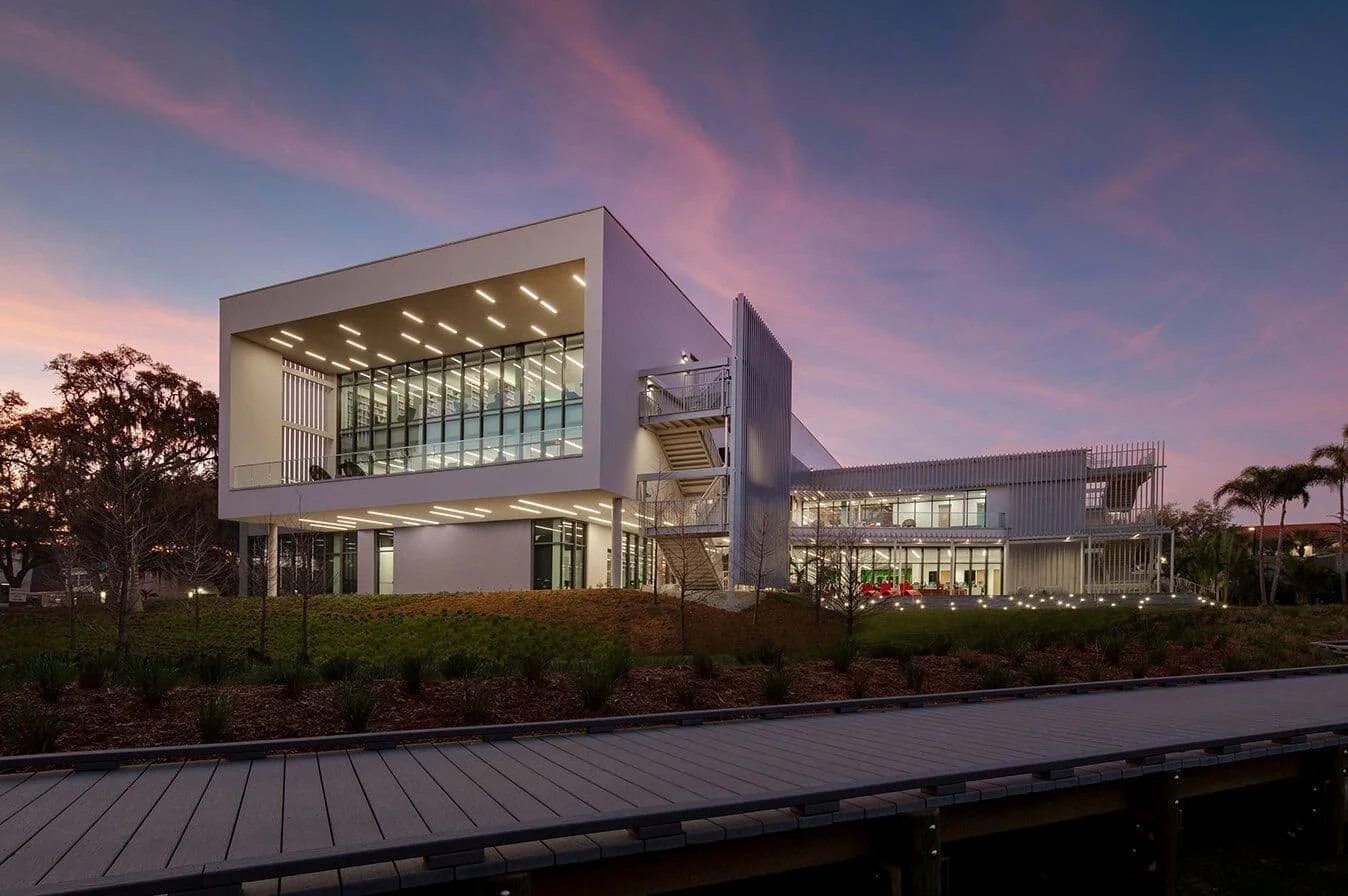 Modern multi-story office building with glass walls and outdoor staircases at sunset, illuminated by interior and exterior lights.