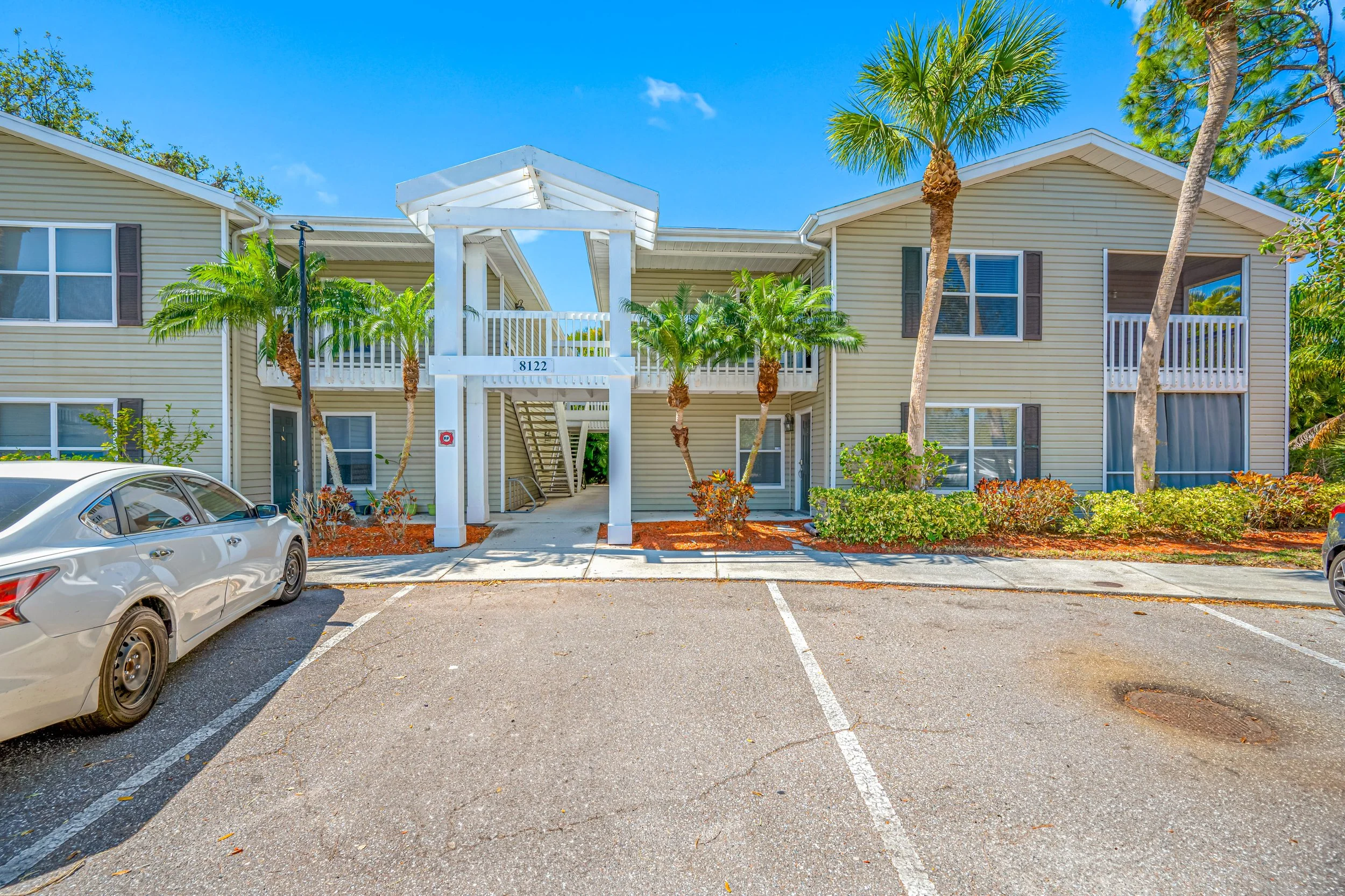 Two-story beige apartment building with palm trees in front and a parking lot with cars.