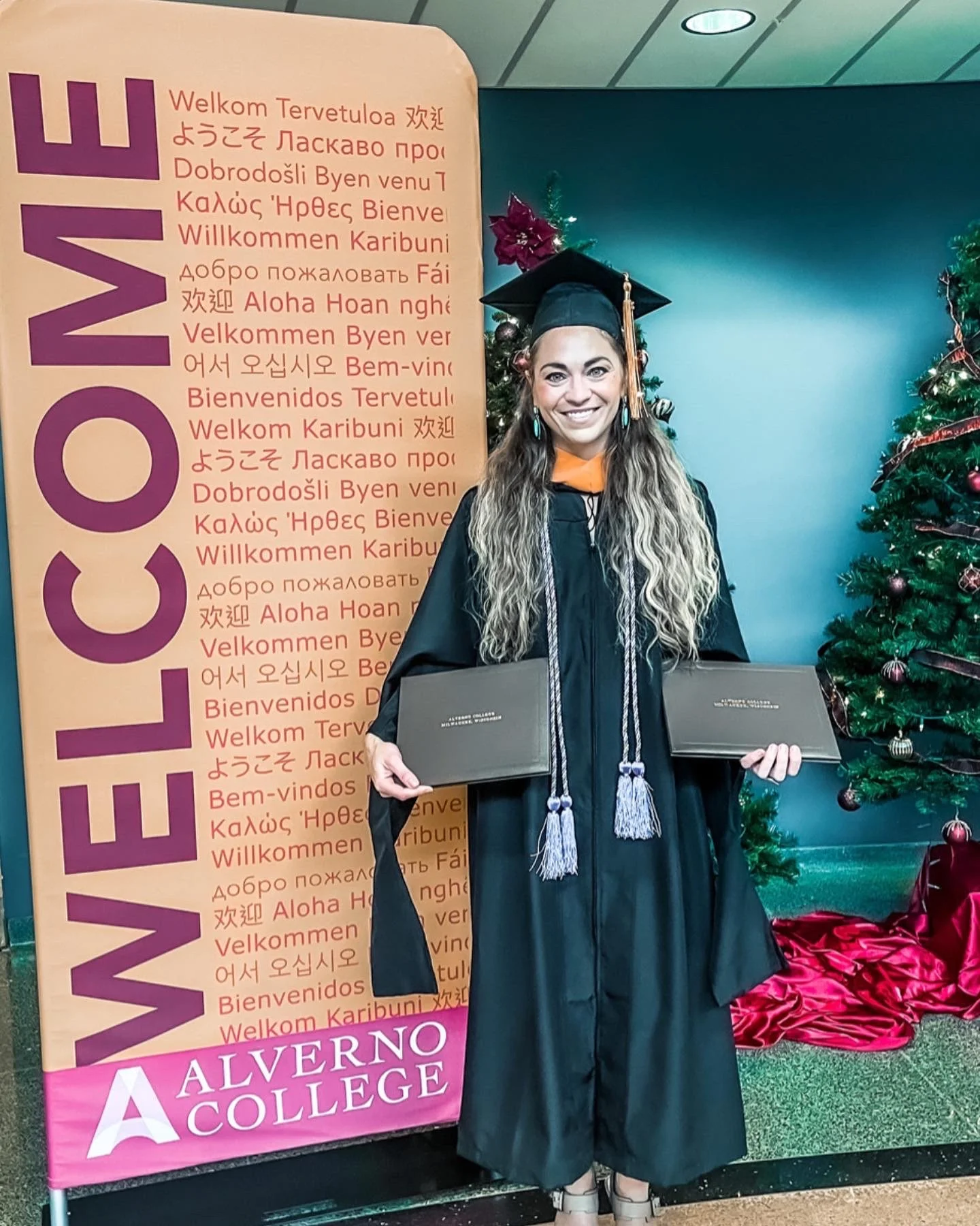 A young woman in graduation cap and gown holding diploma certificates, standing next to a Christmas tree and a large welcome sign with multilingual welcome messages, at Alverno College.
