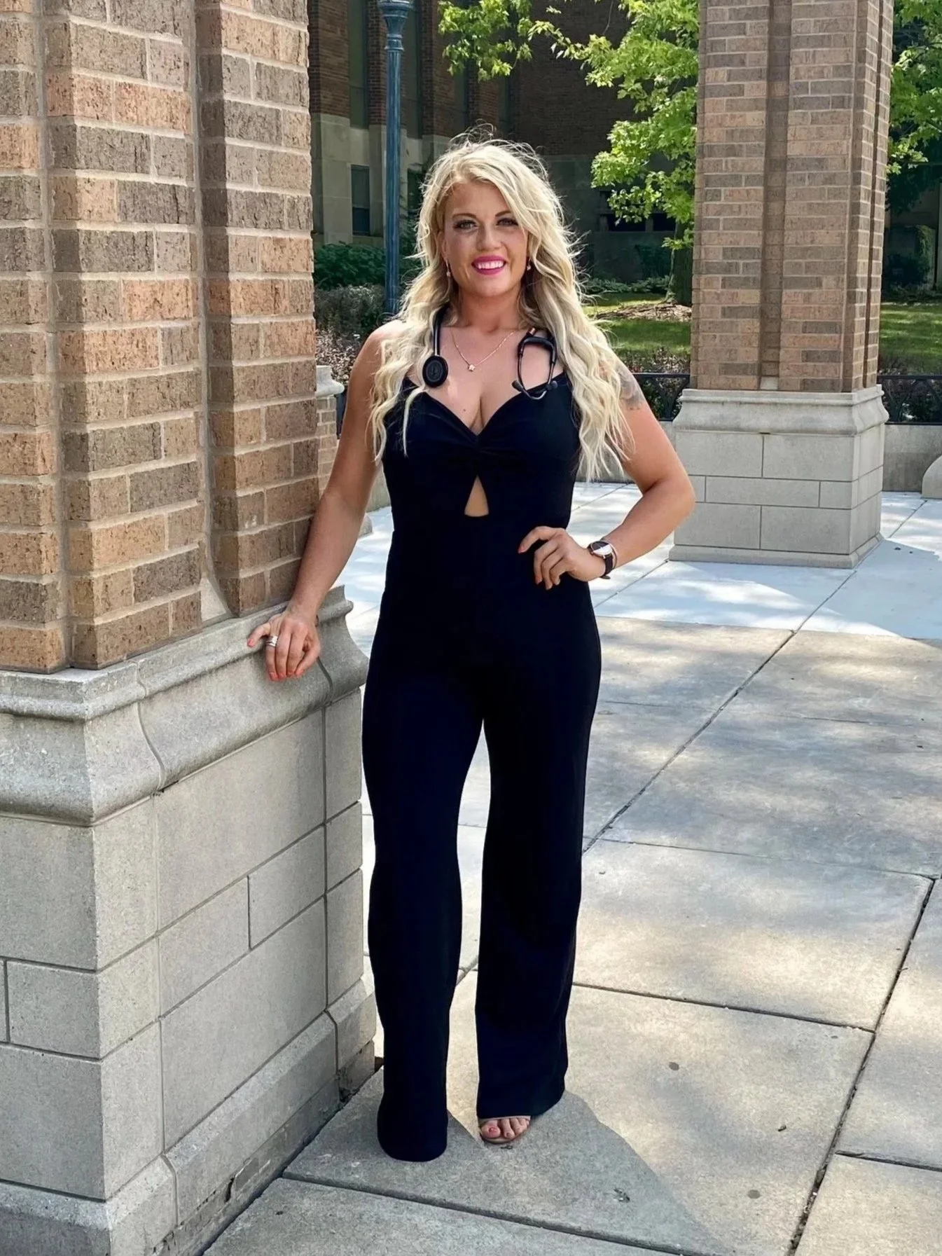 A woman with long blonde hair, wearing a black jumpsuit with a cutout in the front, standing outdoors next to a brick building with green trees in the background. She has a stethoscope around her neck and is smiling.