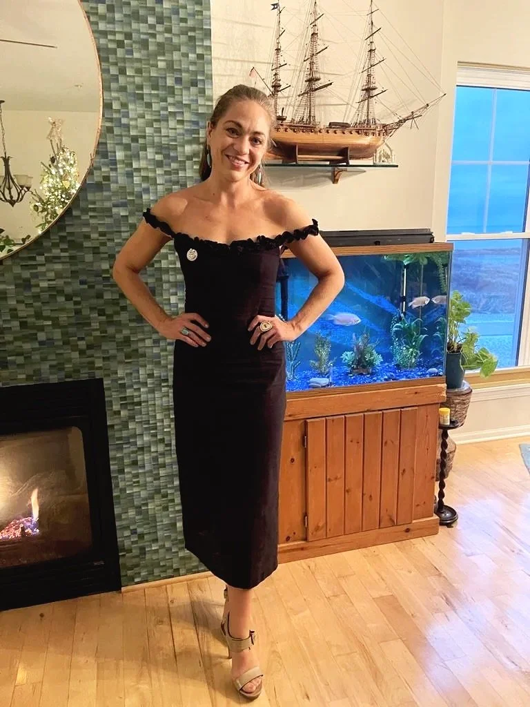 A woman in a black off-shoulder dress and beige heels standing indoors in front of a fish tank, a green textured wall, a mirror, and a model ship on a shelf.
