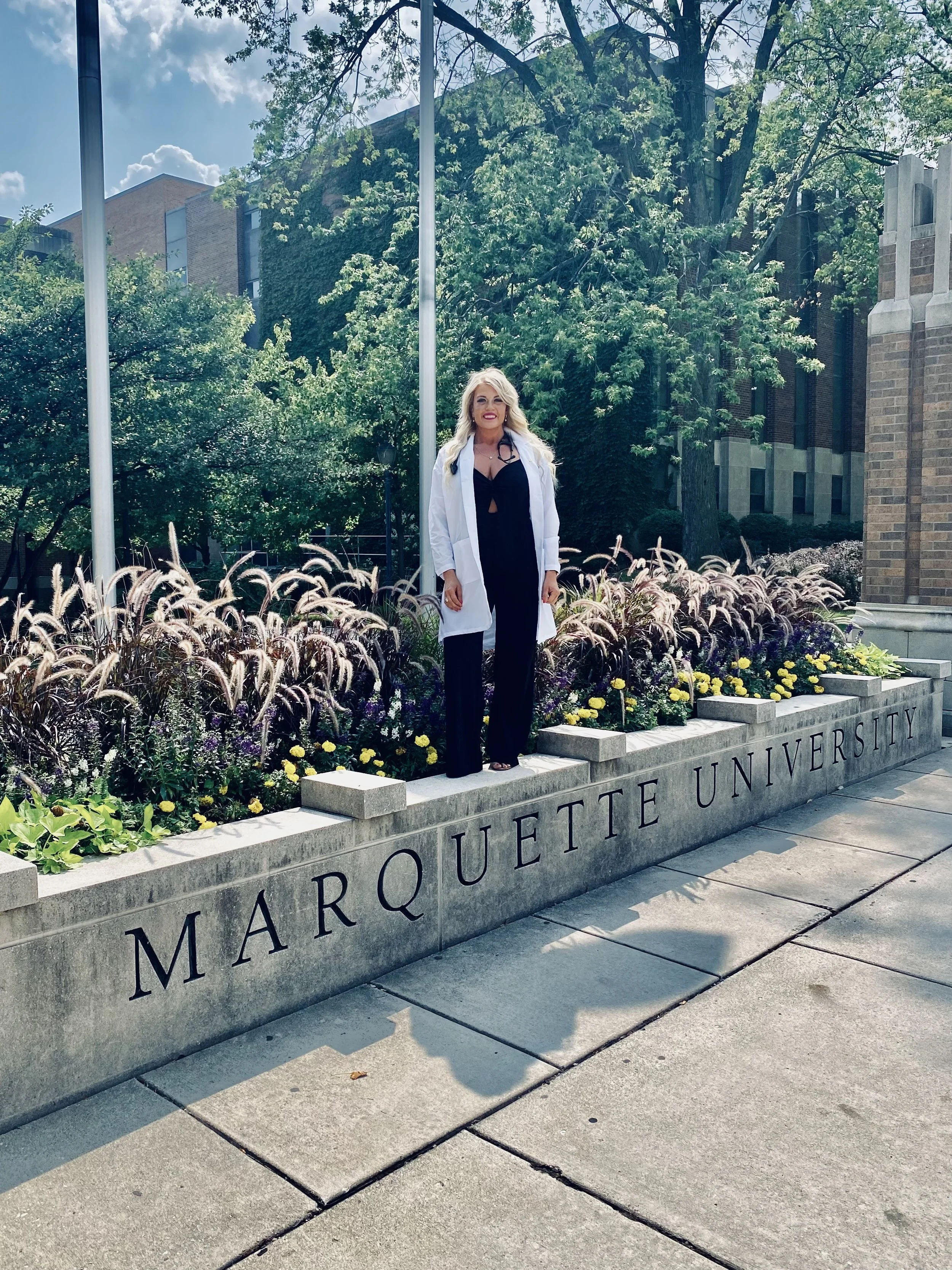 A woman standing on a planter with the words 'Marquette University' engraved on it, surrounded by flowers and greenery, with a university building and trees in the background.