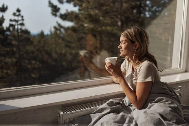 A woman sitting on a bed by a large window, holding a cup and enjoying the sunlight.