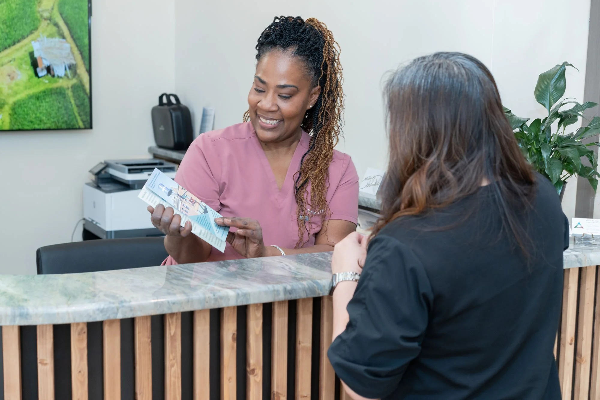A female healthcare professional wearing pink scrubs talking to a woman at a reception desk in an office or clinic setting. The healthcare professional is holding a brochure and smiling. There is a large plant and office equipment in the background.