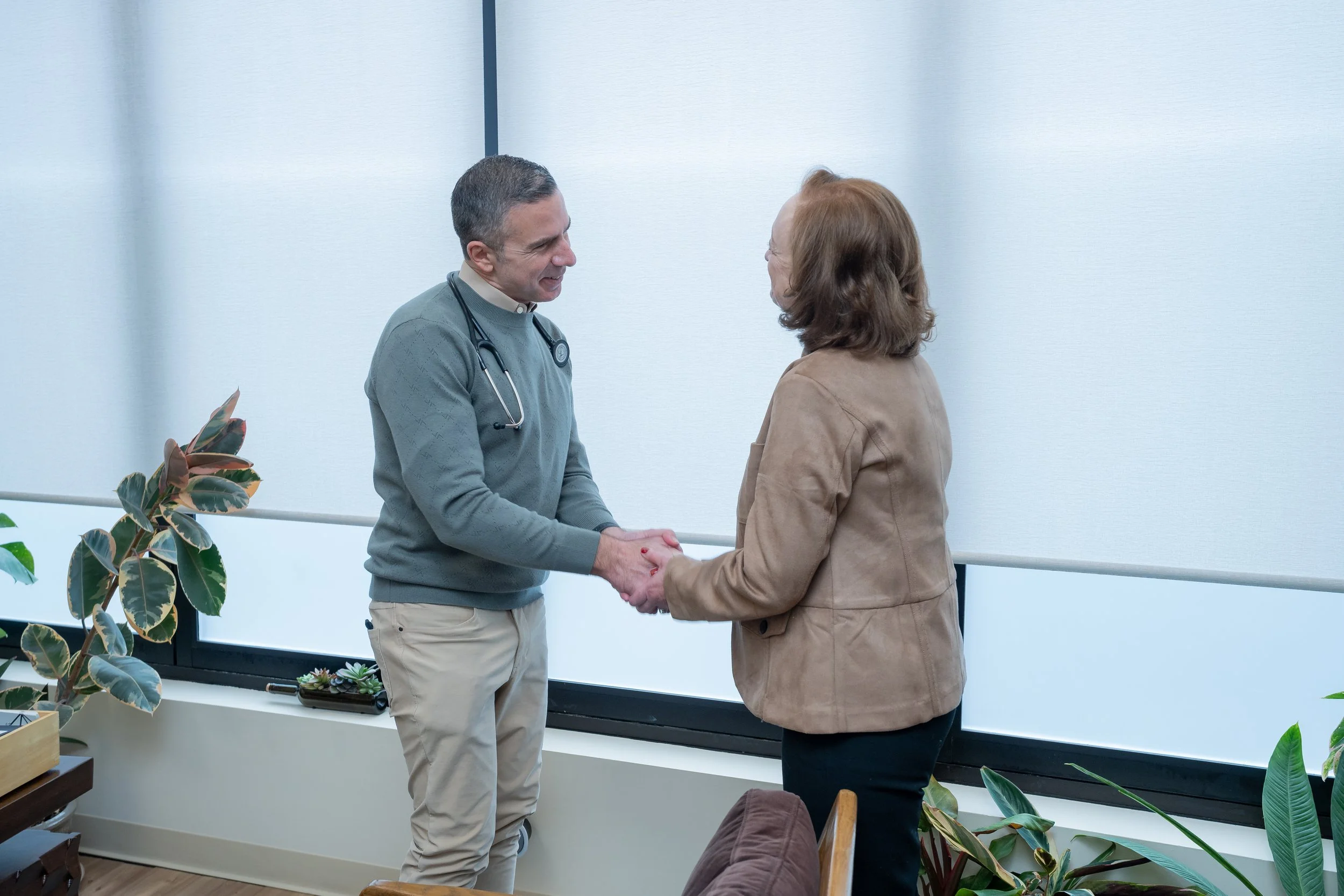 A male doctor shaking hands with a woman in a medical office, both smiling and making eye contact, with houseplants nearby.