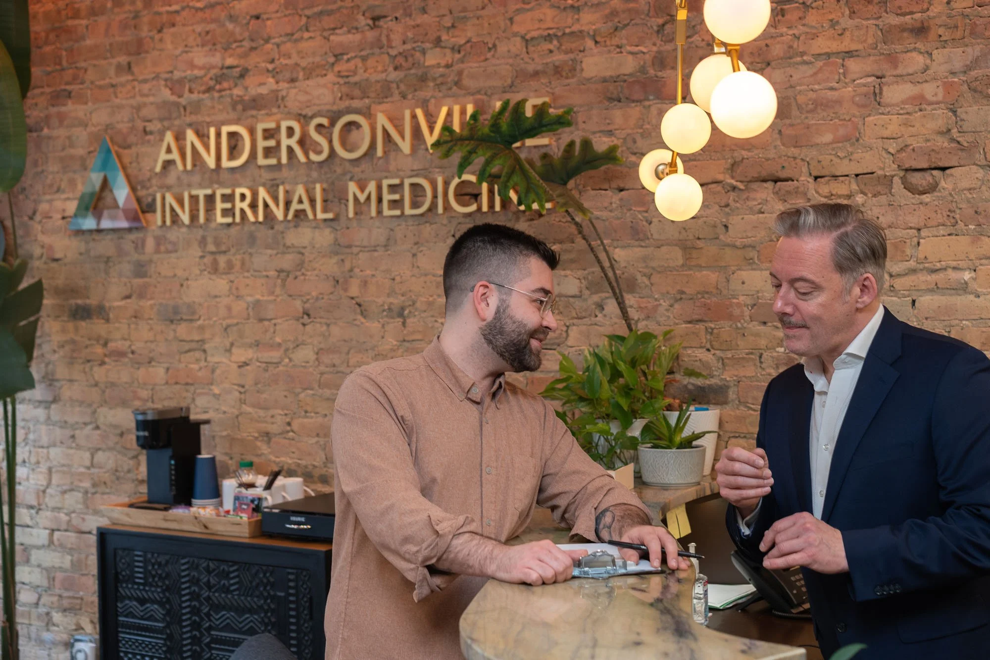 Two men are working together at a reception desk in an office with a brick wall background. A sign on the wall reads 'Andersonville Internal Medicine'.