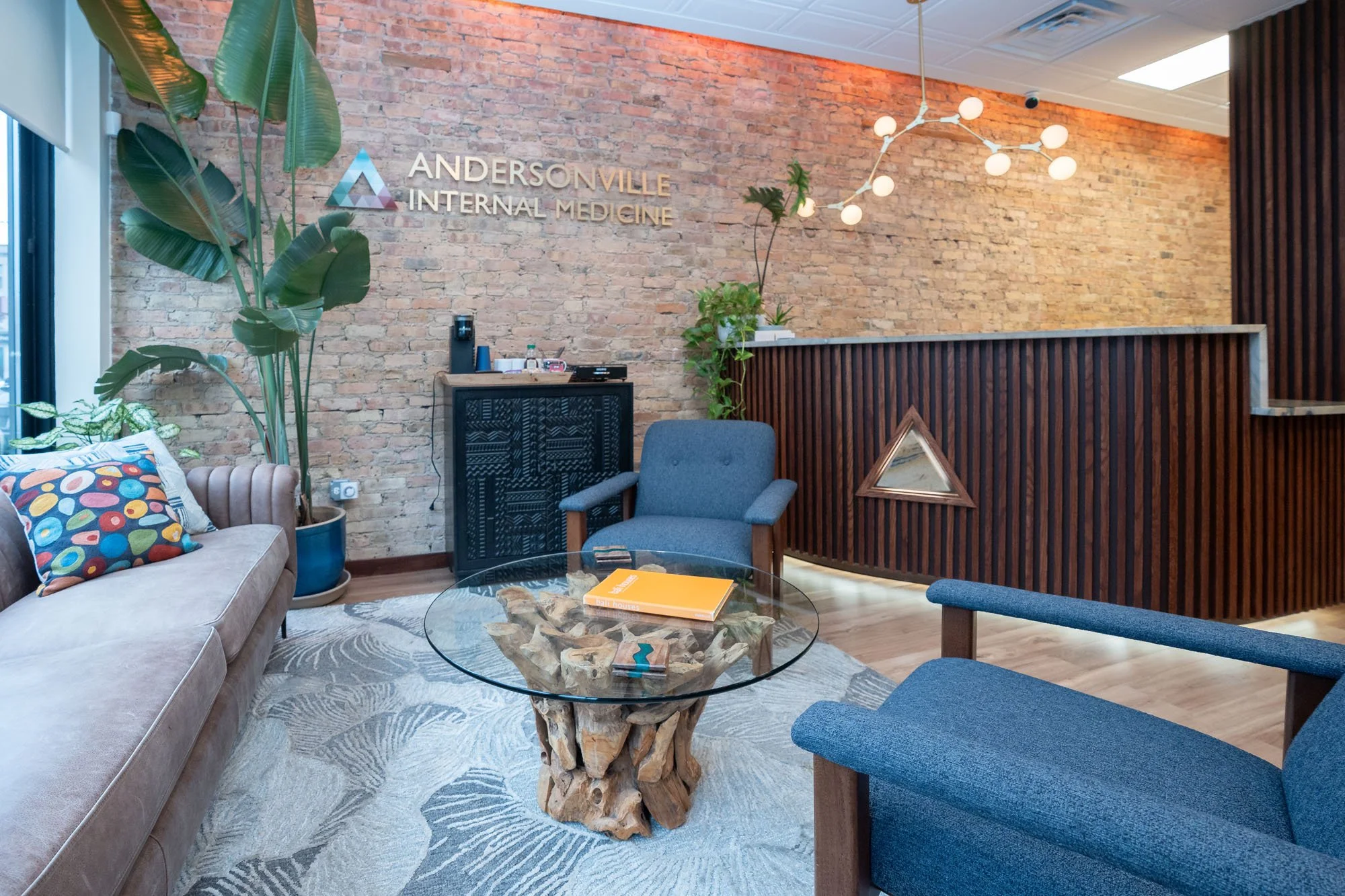 Indoor office reception area with exposed brick wall, Andersonville Internal Medicine sign, various plants, seating area with beige sofa and blue chairs, glass coffee table, modern chandelier, and decorative wood paneling.