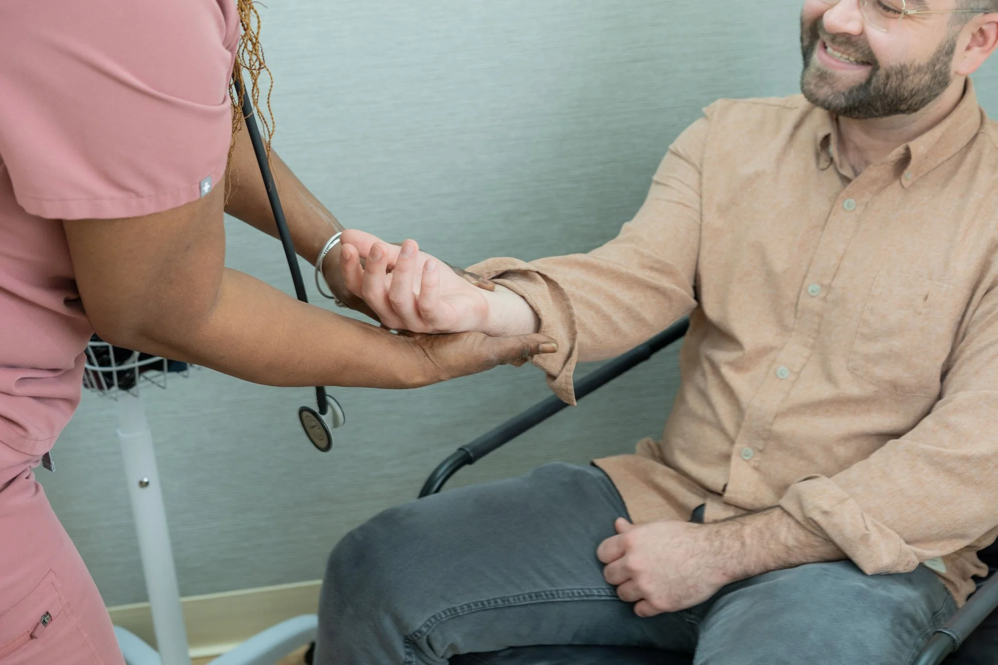 A healthcare worker in pink scrubs is checking a man's blood pressure using a stethoscope and blood pressure cuff.