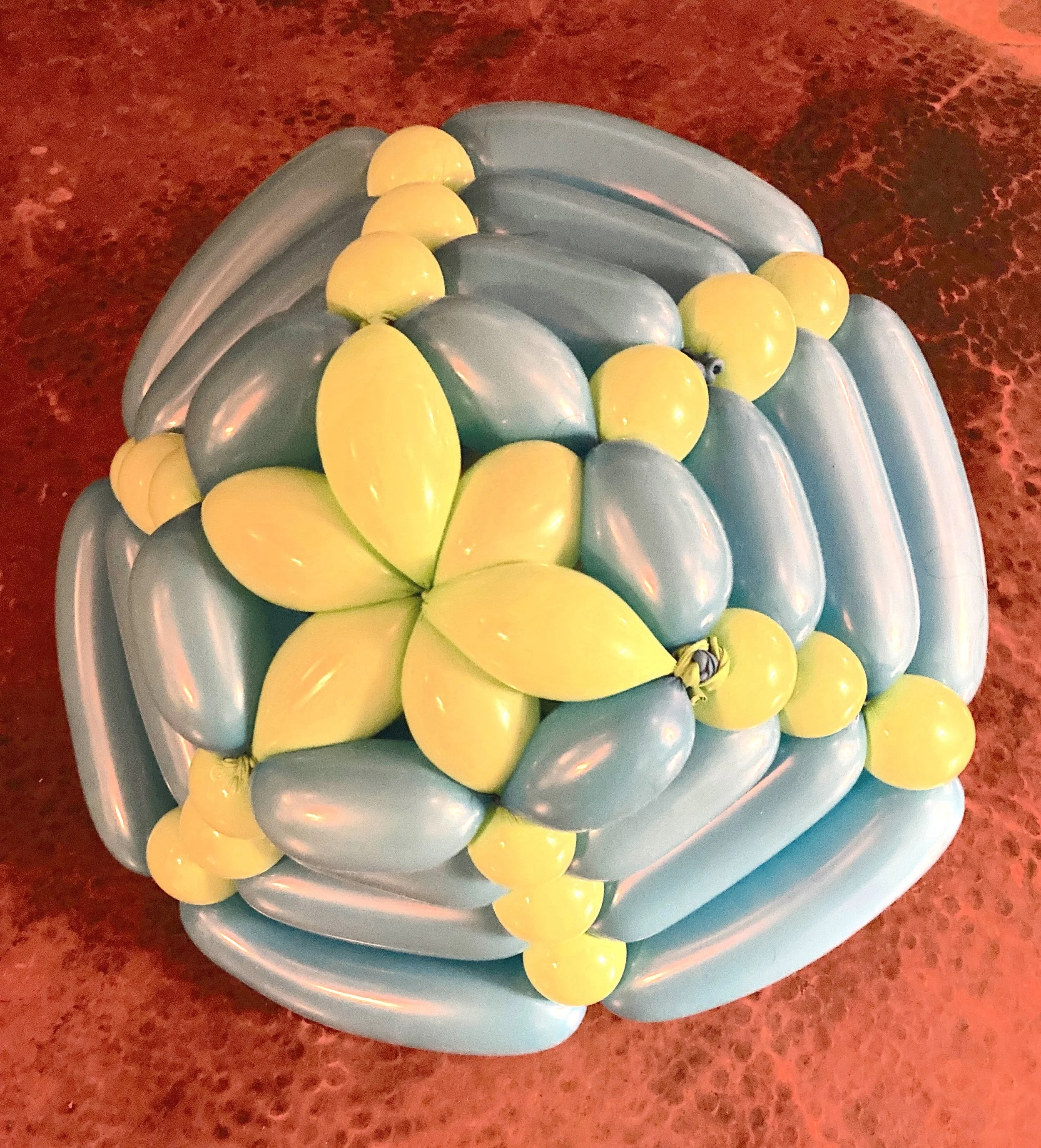 Colorful balloon sculpture resembling a cake with yellow and blue balloons arranged in a floral pattern on a red textured surface. (Created by O.E. Zelmanovich. Zoe Zelmanovich. Zoe Mizuho)