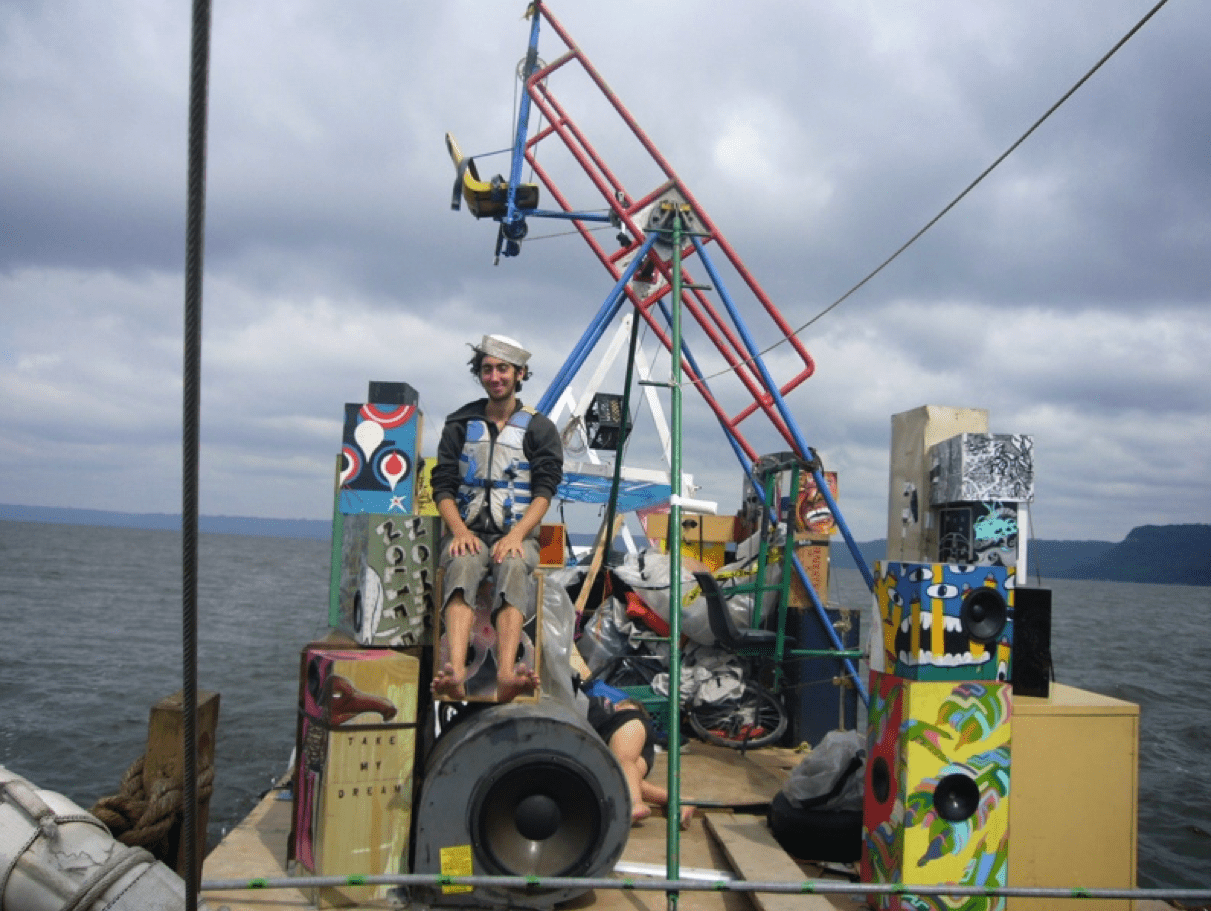 A man sitting on a colorful, makeshift raft decorated with speakers, painted boxes, and various objects on a body of water with a cloudy sky in the background.
