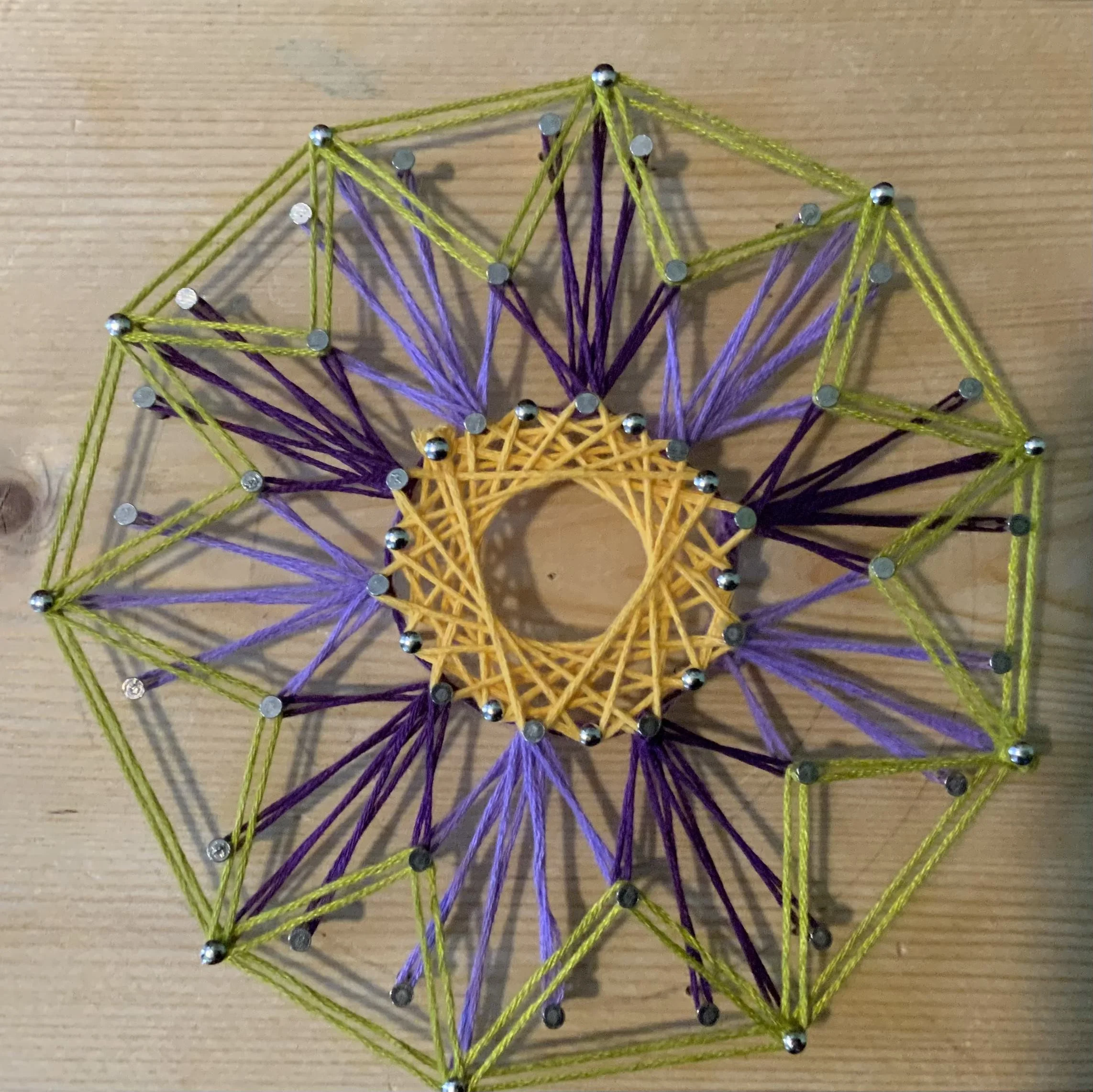 Colorful string art with yellow, purple, and green strings arranged in a geometric pattern on a wooden surface.  (Created by O.E. Zelmanovich. Zoe Zelmanovich. Zoe Mizuho)