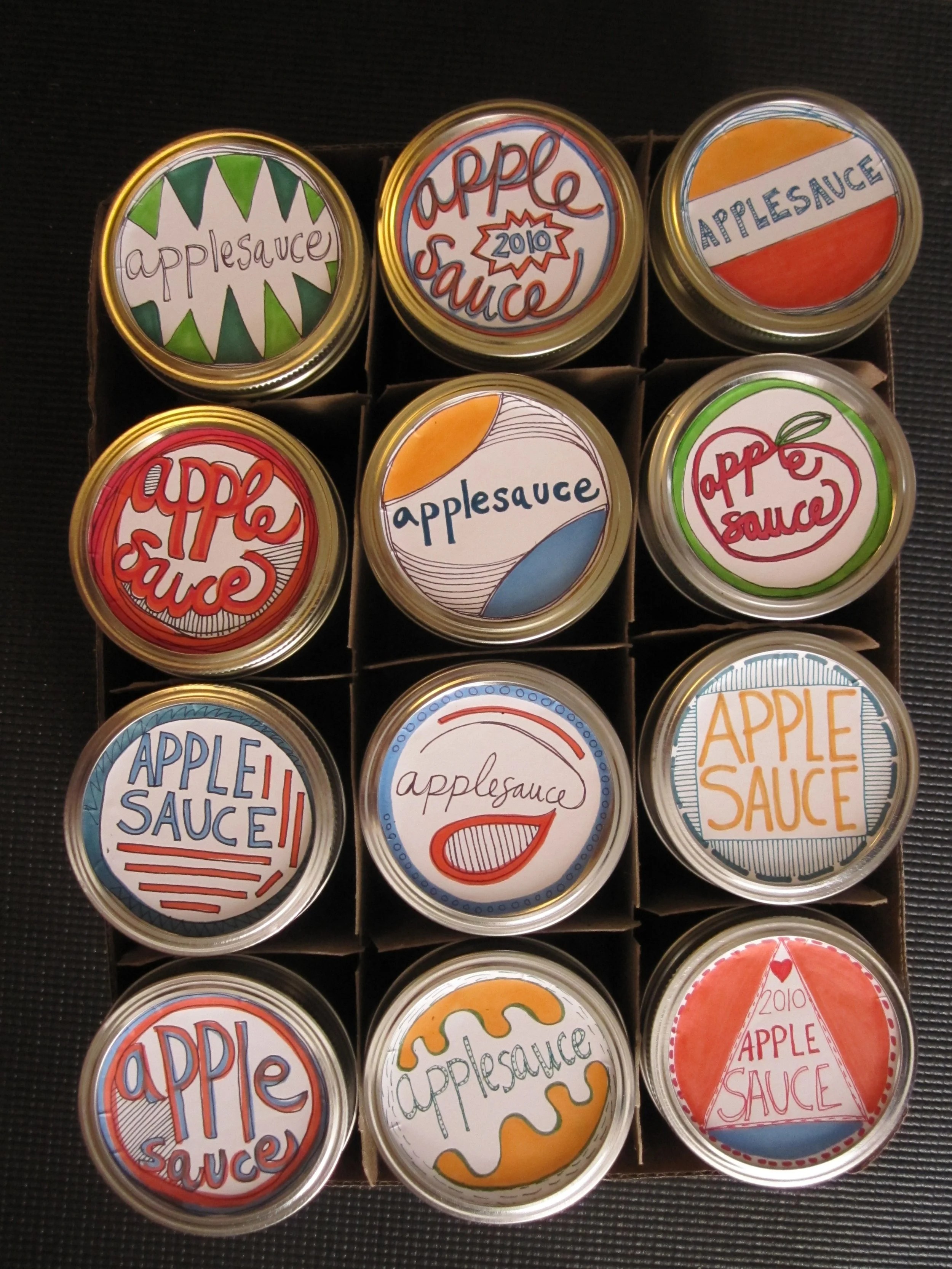 A collection of twelve jars of apple sauce, each with hand-drawn labels and designs, arranged in a 3x4 grid inside a cardboard holder. (Created by O.E. Zelmanovich. Zoe Zelmanovich. Zoe Mizuho)