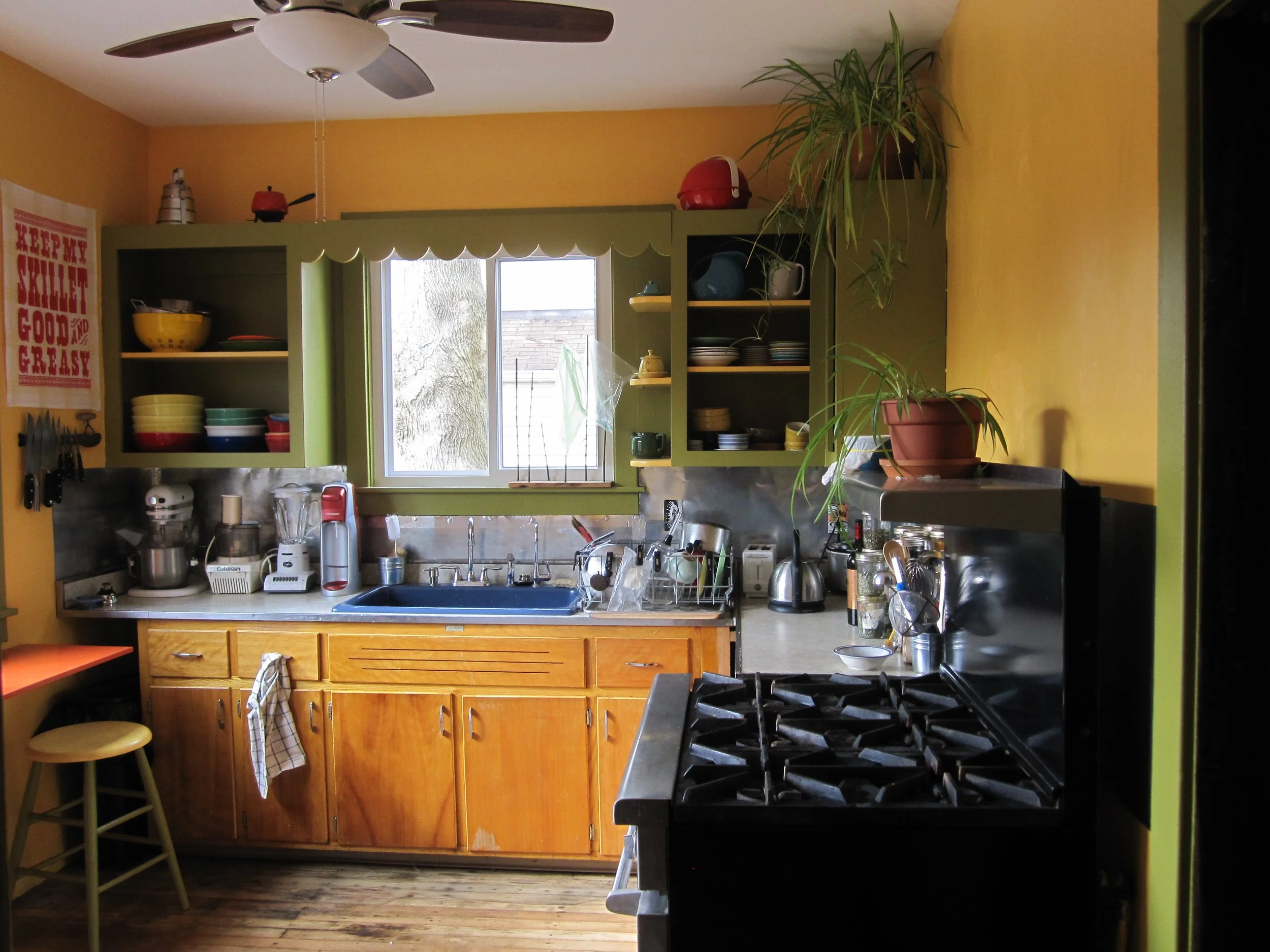 A colorful kitchen with yellow walls, green cabinets, a window above the sink, and a black stove. Various kitchen appliances, dishes, and plants are visible.  (Designed by O.E. Zelmanovich. Zoe Zelmanovich. Zoe Mizuho)