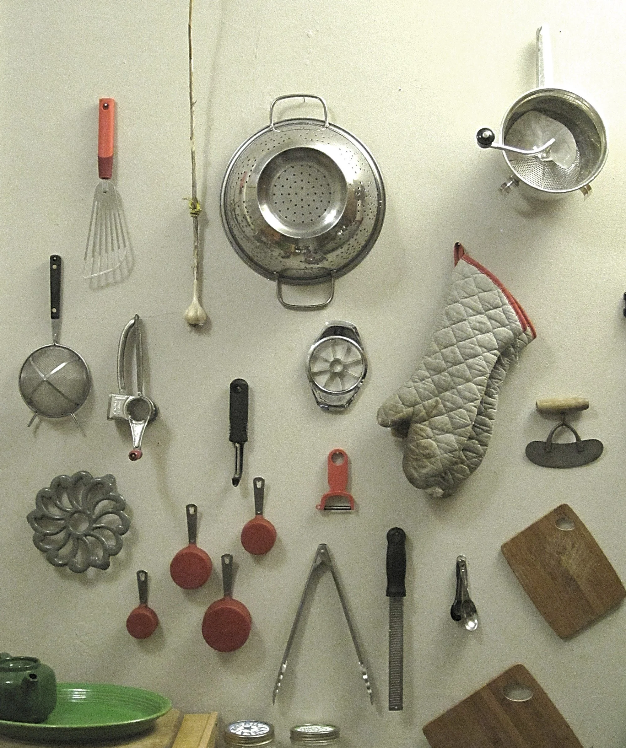 Kitchen wall with hanging utensils including a whisk, garlic, colander, ladle, measuring cups, cutting boards, and a quilted oven mitt.  (Designed by O.E. Zelmanovich. Zoe Zelmanovich. Zoe Mizuho)