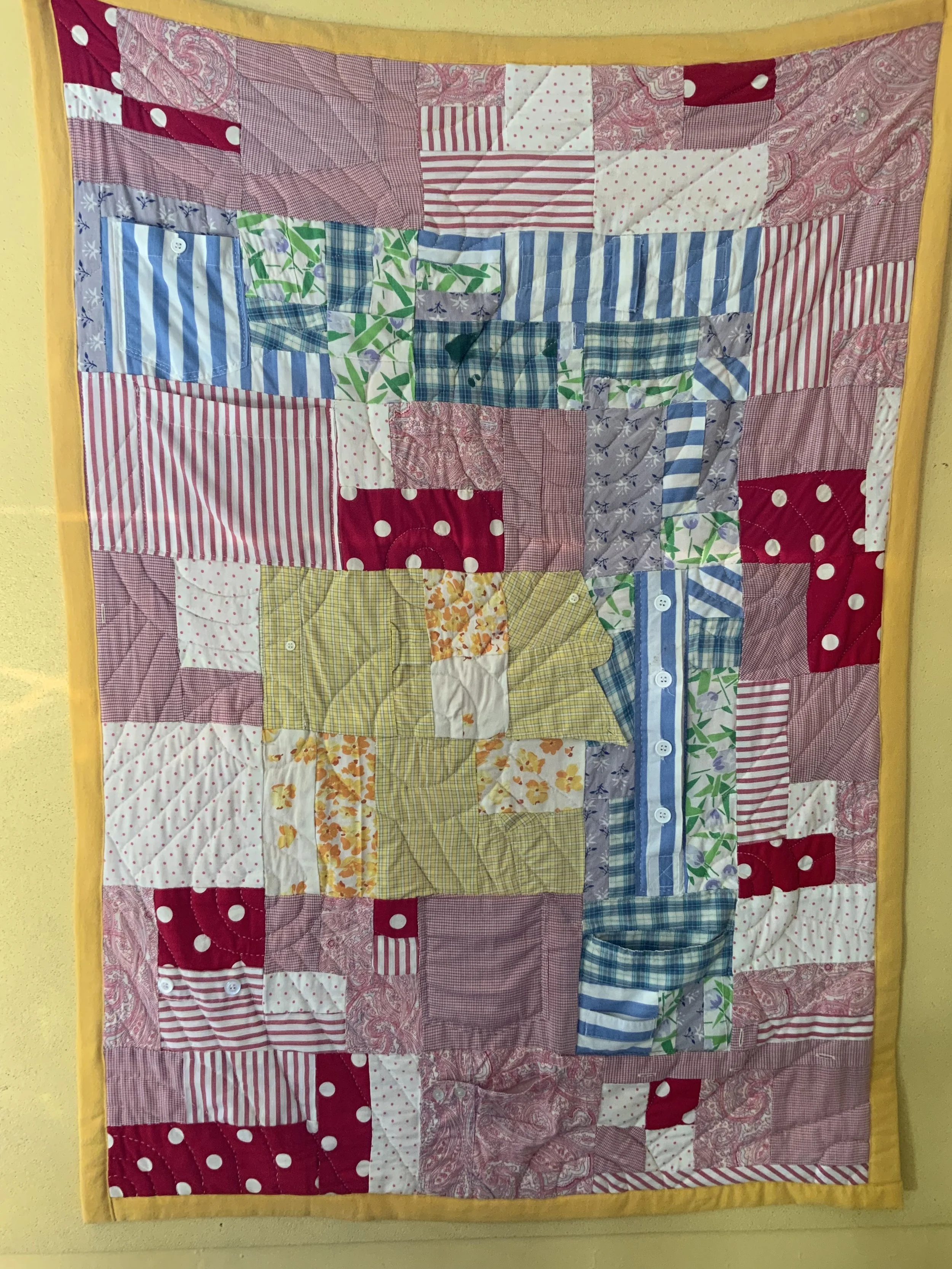 A colorful patchwork quilt with various patterns of red, pink, yellow, blue, green, and white fabrics, bordered with yellow fabric. (Created by O.E. Zelmanovich. Zoe Zelmanovich. Zoe Mizuho)