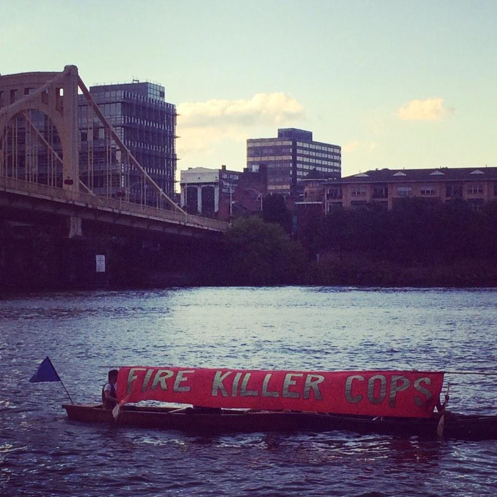 Zoe and friends in a canoe with the banner 'FIRE KILLER COPS' on the Allegheny River in Pittsburgh during the Fraternal Order of Police (FOP) convention in 2015.